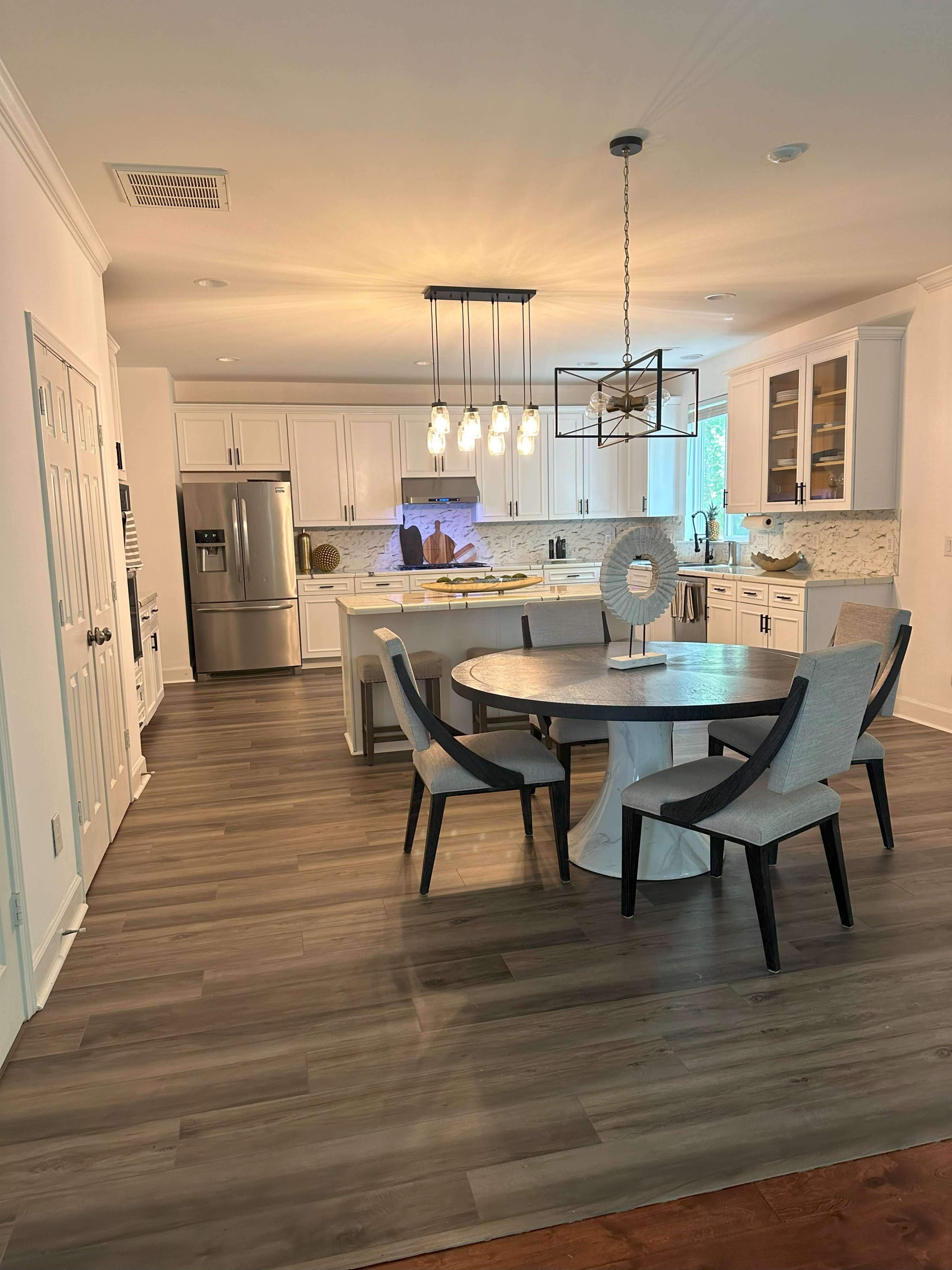 A modern kitchen with a circular dining table and six chairs, featuring white cabinetry, stainless steel appliances, and a pendant light fixture.