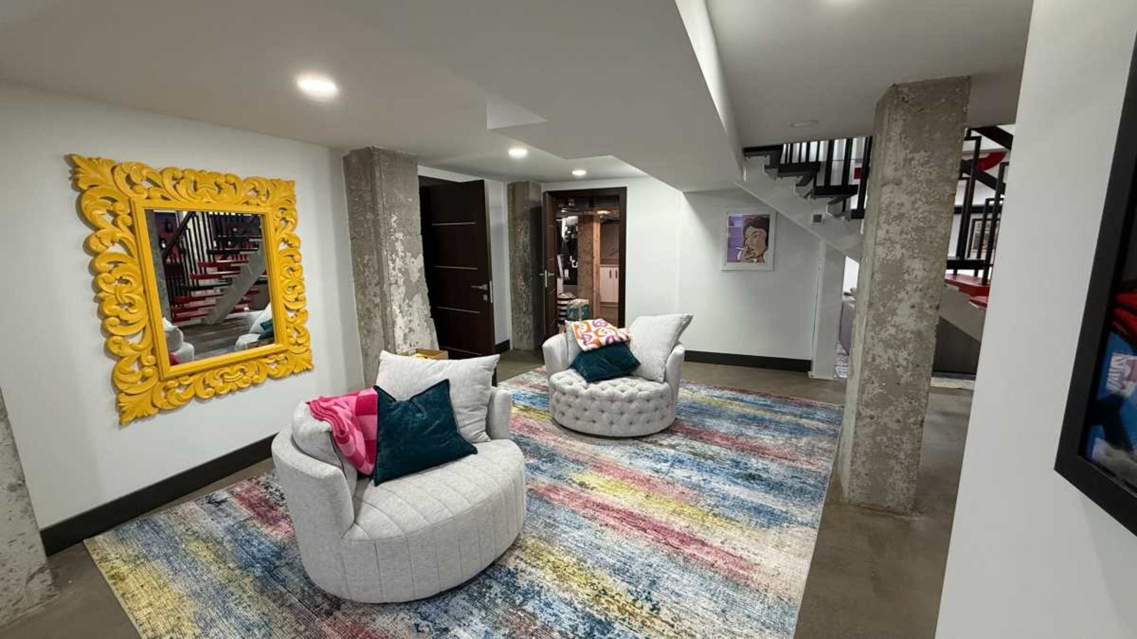 The image shows a basement area featuring two upholstered armchairs, a colorful area rug, and a large ornate mirror on one wall.