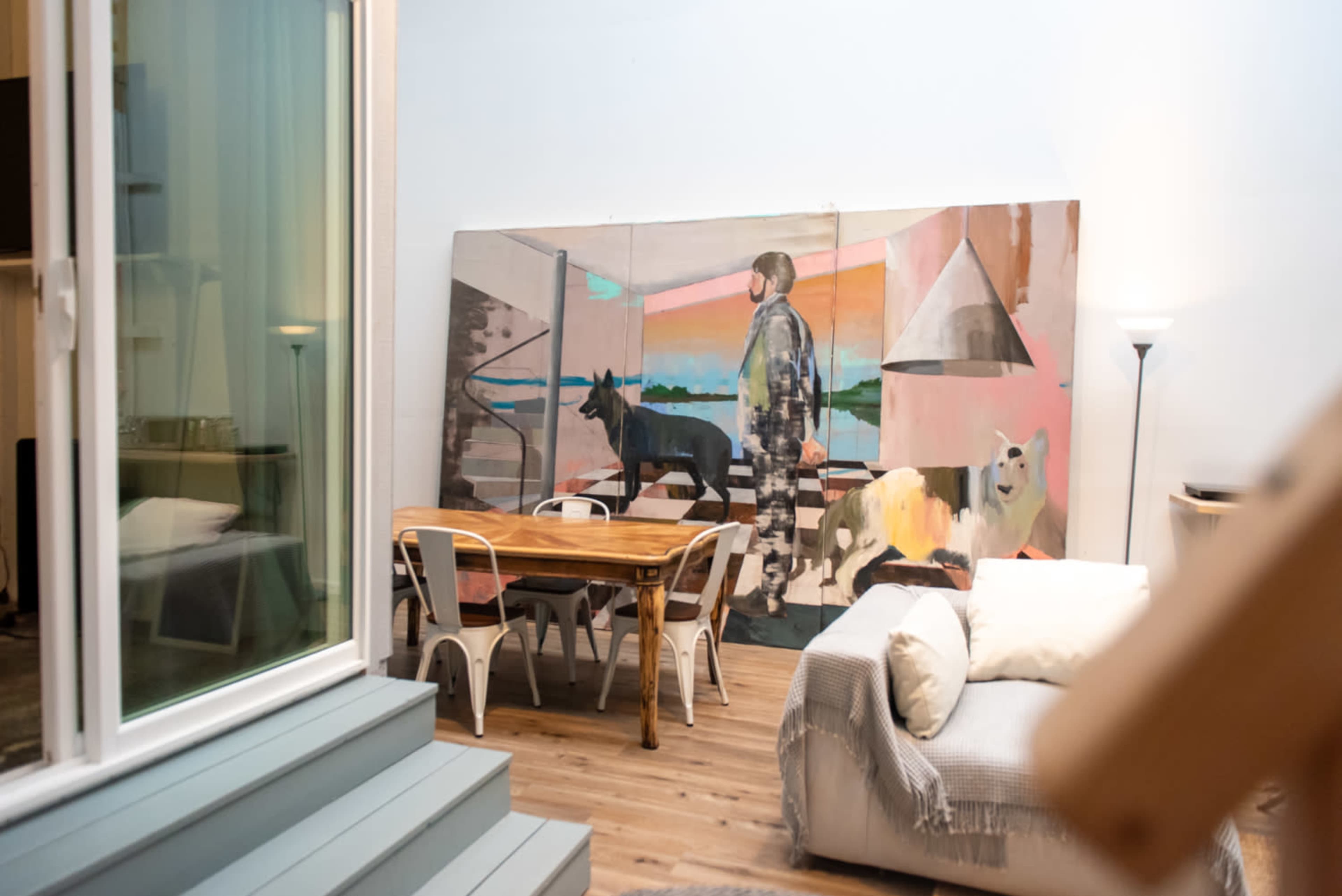 A spacious room features a dining table surrounded by chairs, a couch, and a large mural depicting a figure with dogs.