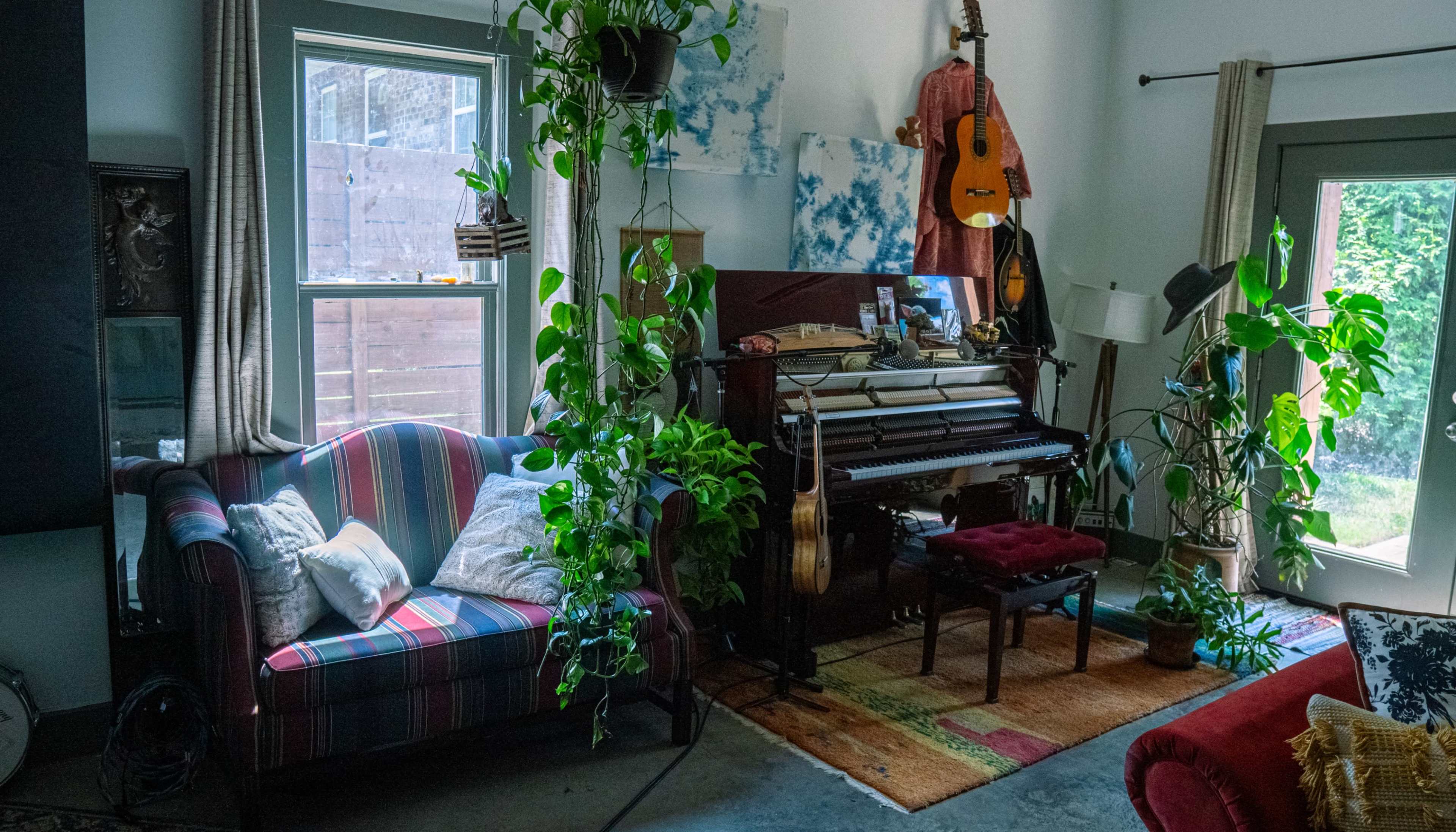 The image shows a cozy living room with a striped sofa, a piano, and plants cascading from shelves and windows.