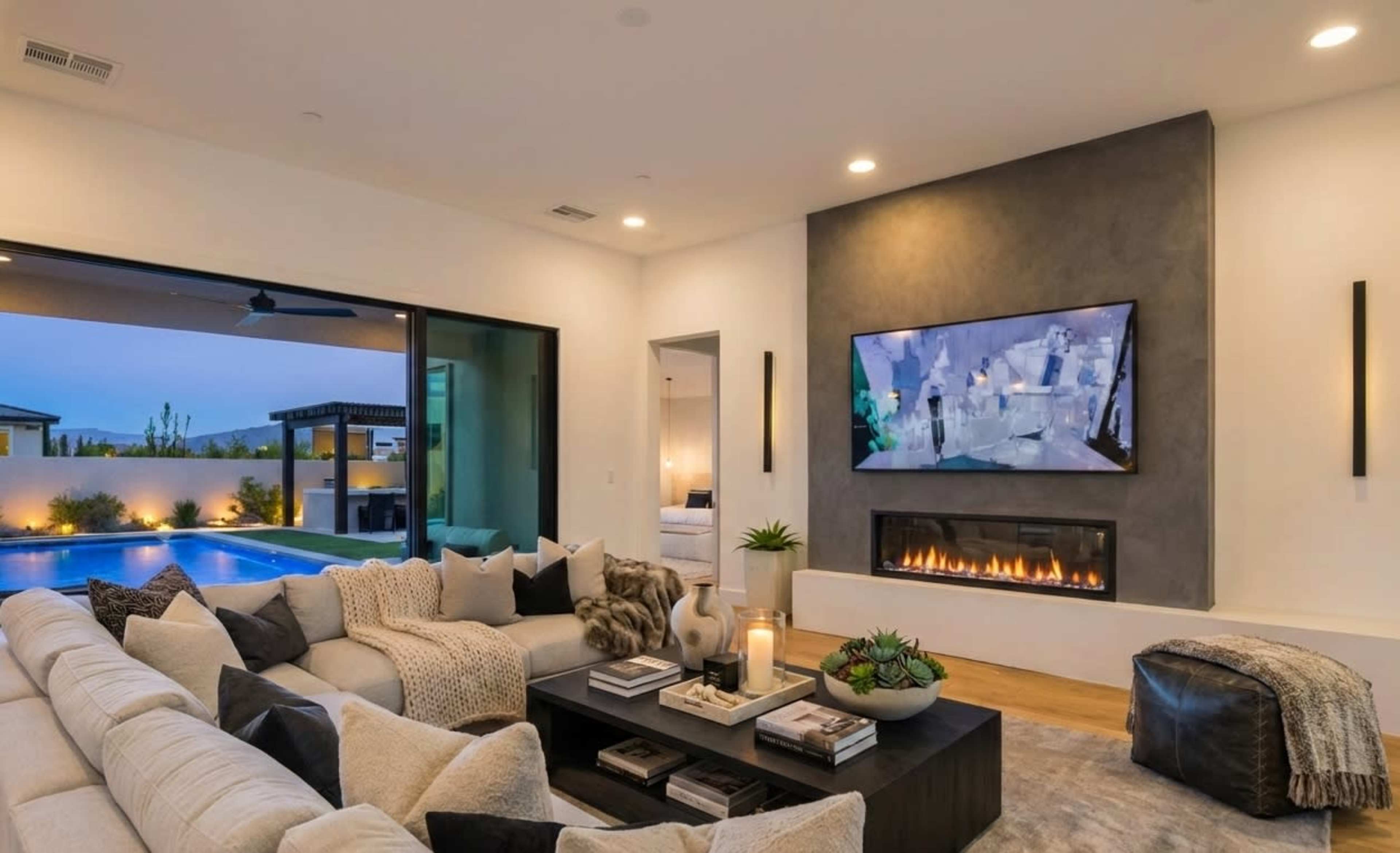 The image shows a modern living room with a large sectional sofa facing a fireplace and a flat-screen television, and glass doors that open to a pool area outside.