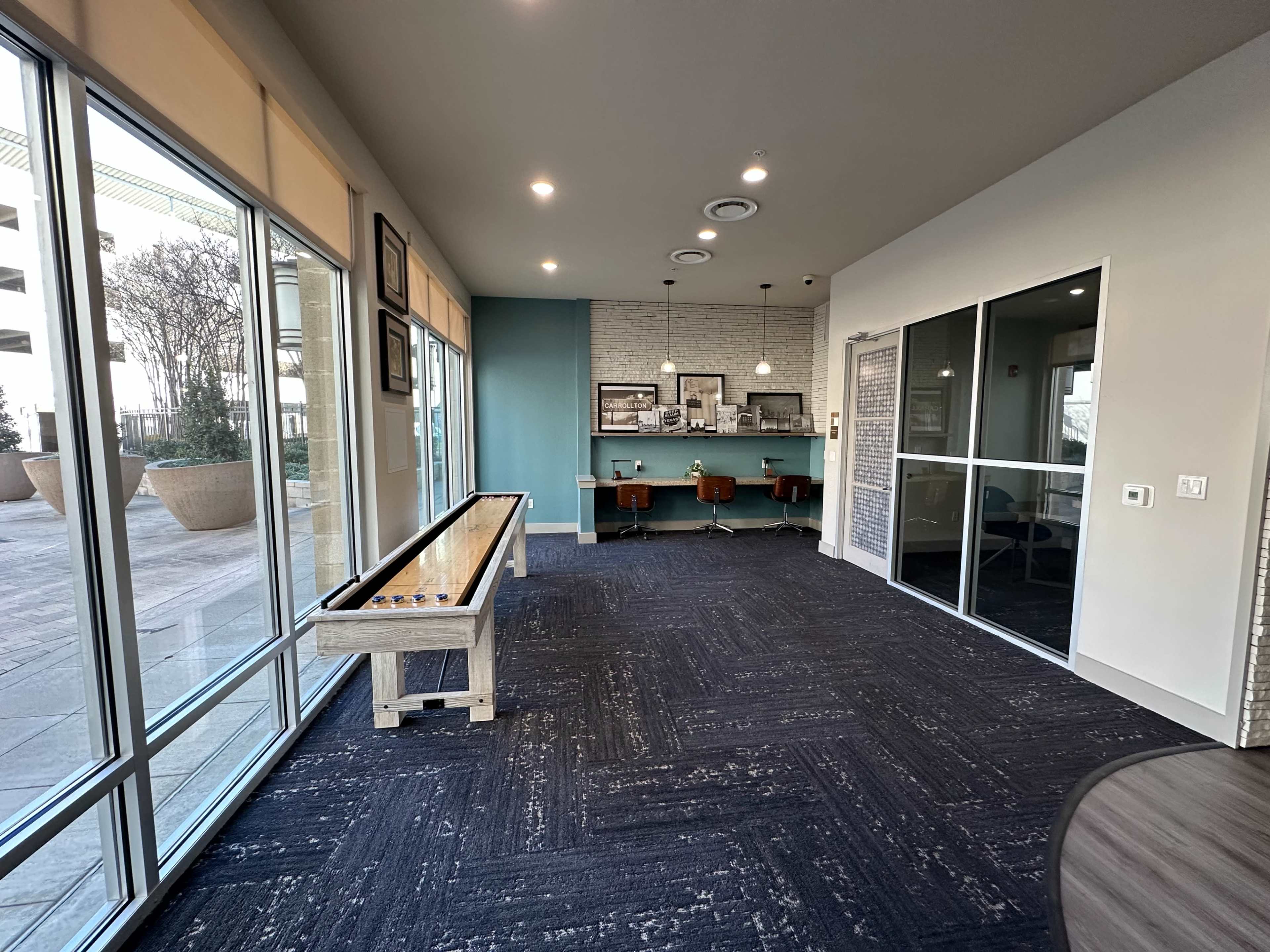 The image shows a modern room with large windows, a shuffleboard table on the left, and a small seating area with bar stools against a blue accent wall.