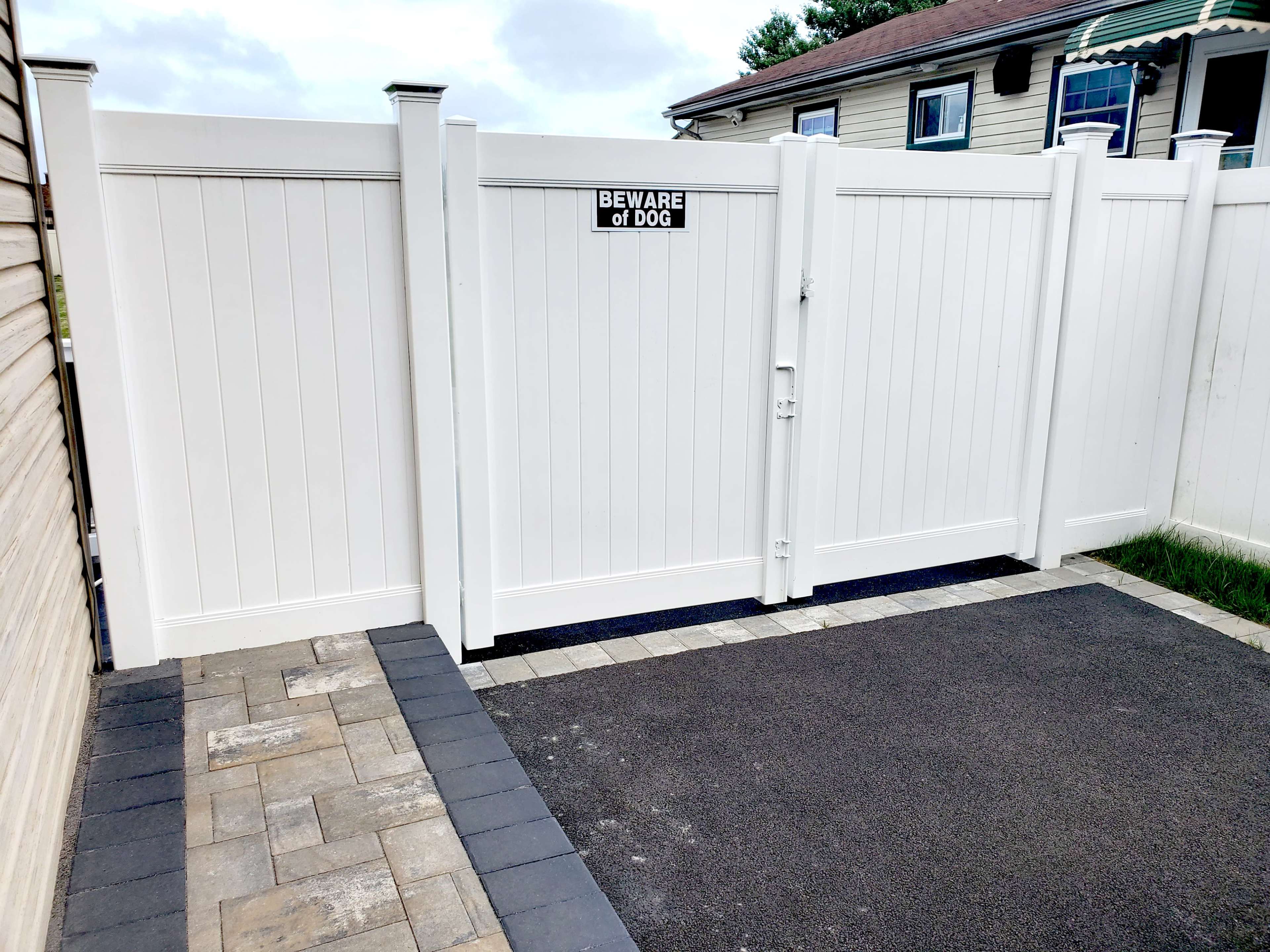 A white vinyl gate with a "Beware of Dog" sign is installed in a fenced area next to a paved driveway.
