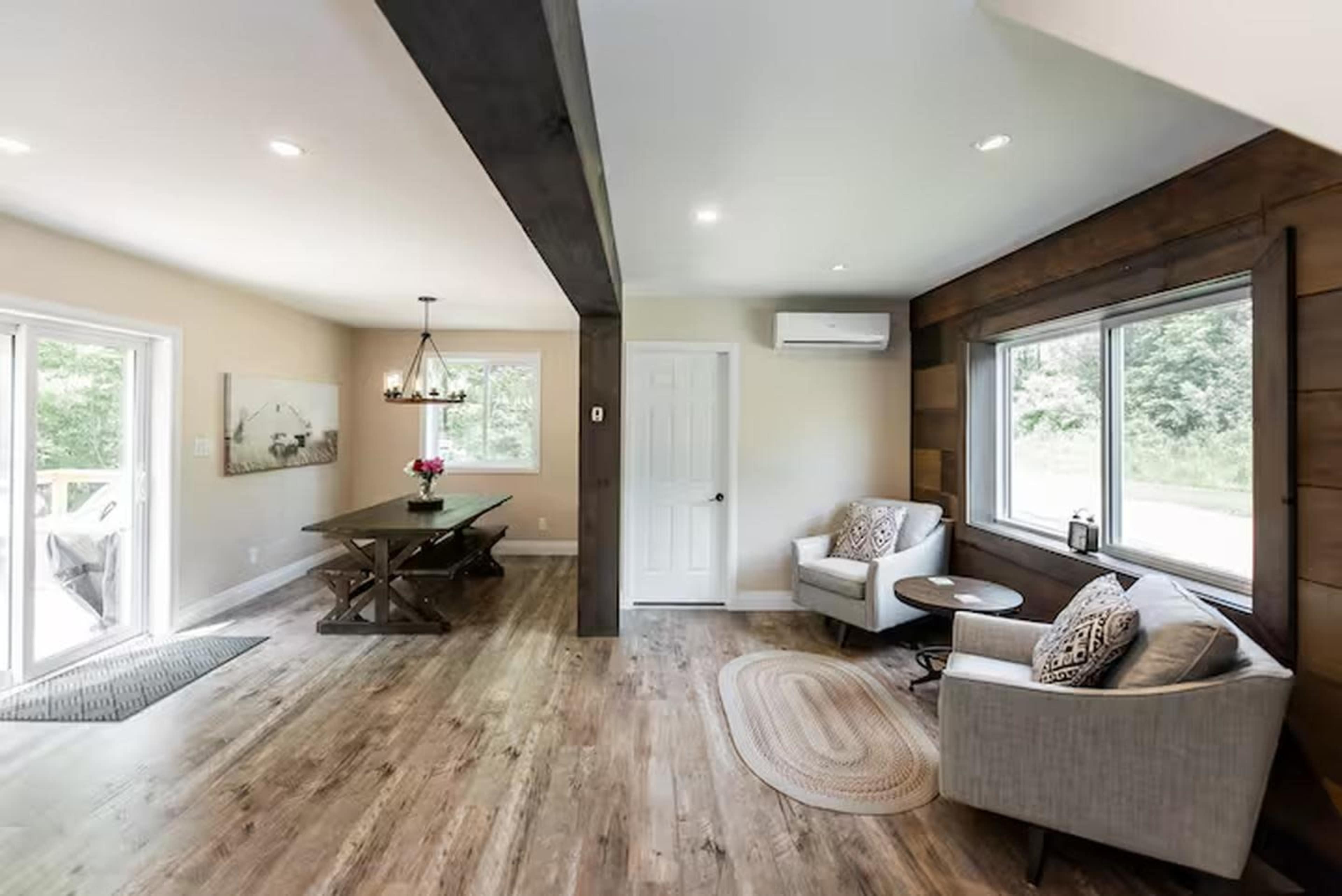 The image shows a modern living space with a dining area, featuring a wooden table, two armchairs, and large windows that allow natural light to enter.