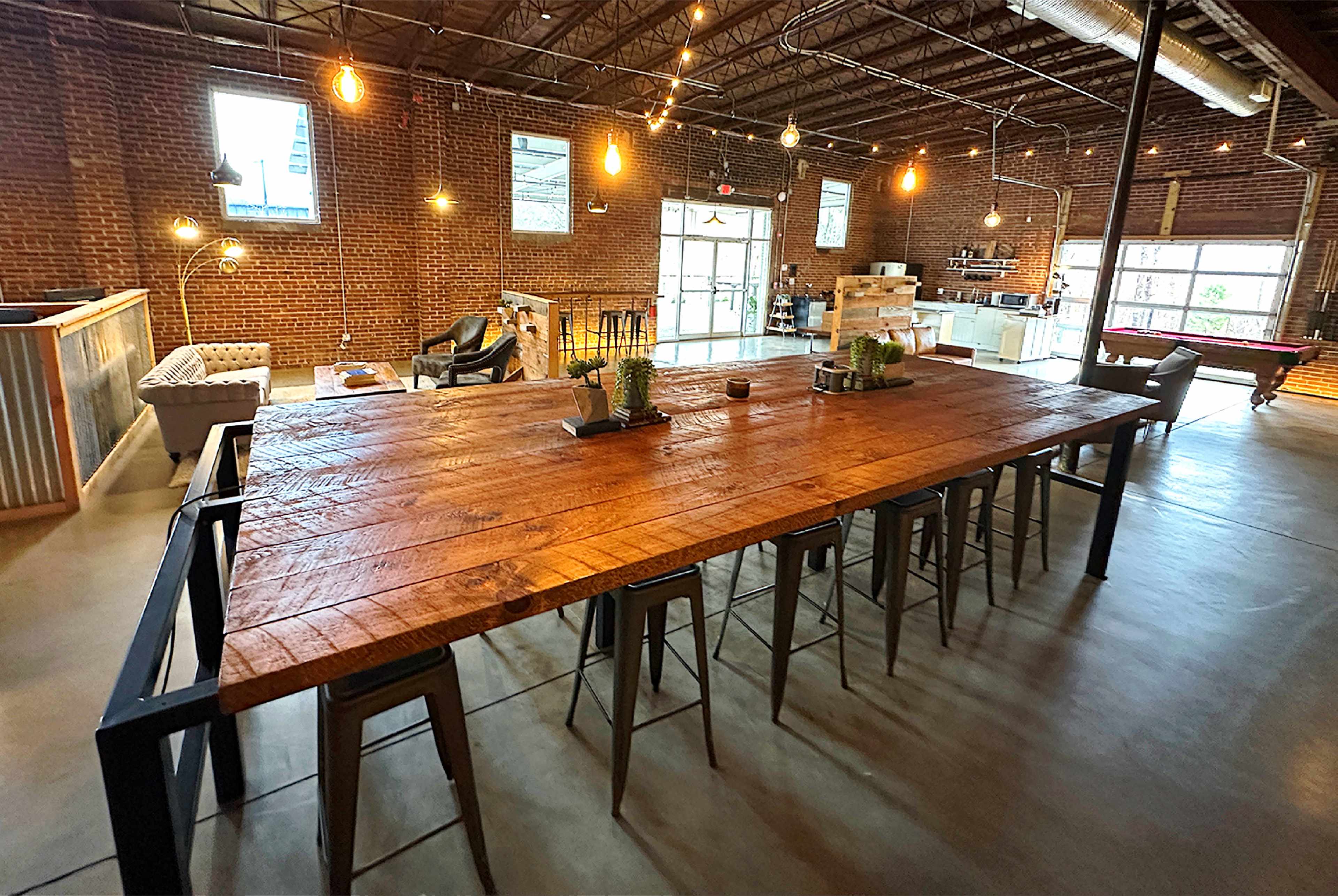 A large wooden table with metal stools is positioned in a spacious, industrial-style room with exposed brick walls and hanging light fixtures.