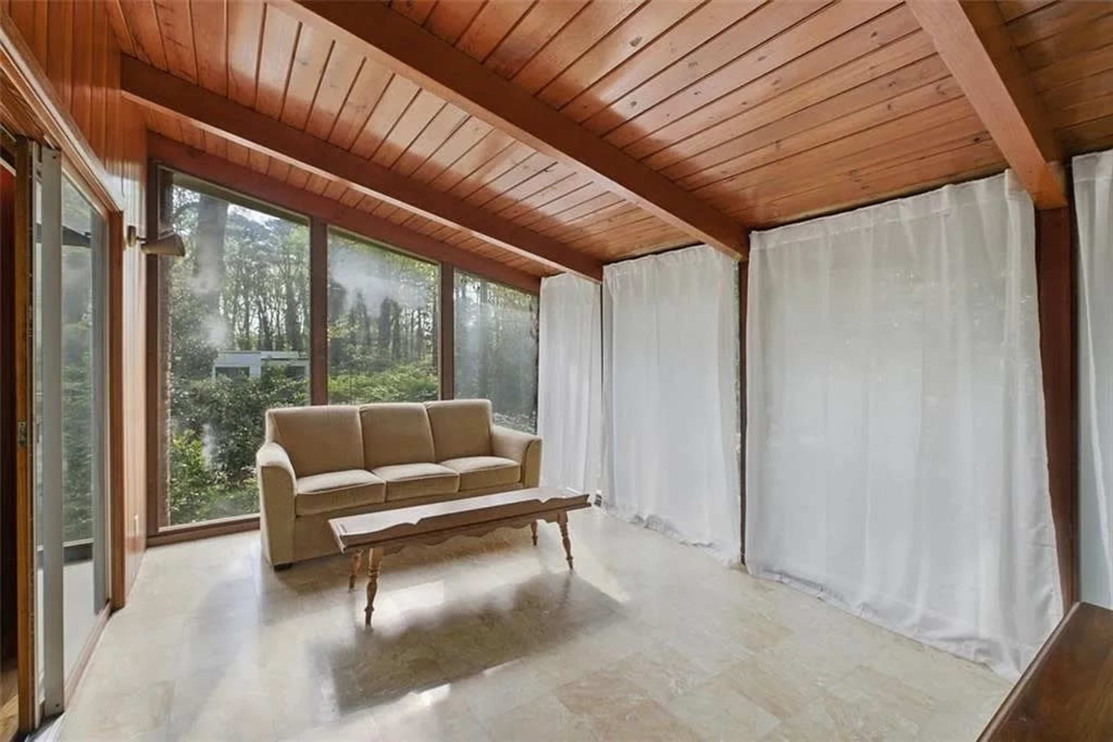 The image shows a sunlit room with large windows, a beige couch, and a wooden coffee table on a tiled floor.
