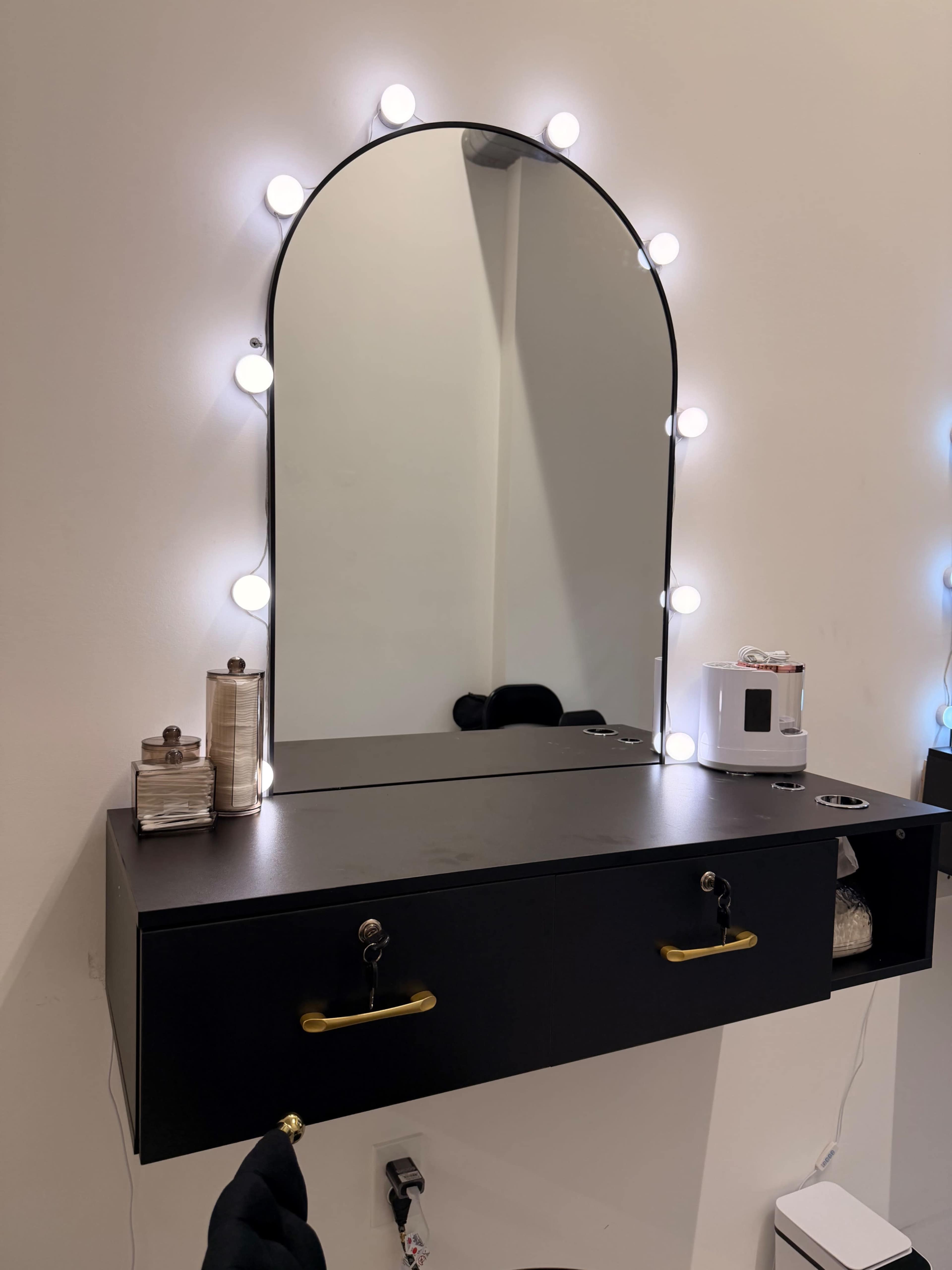 The image shows a wall-mounted black vanity with a large arched mirror surrounded by round light bulbs, featuring two drawers below and a small appliance beside it.