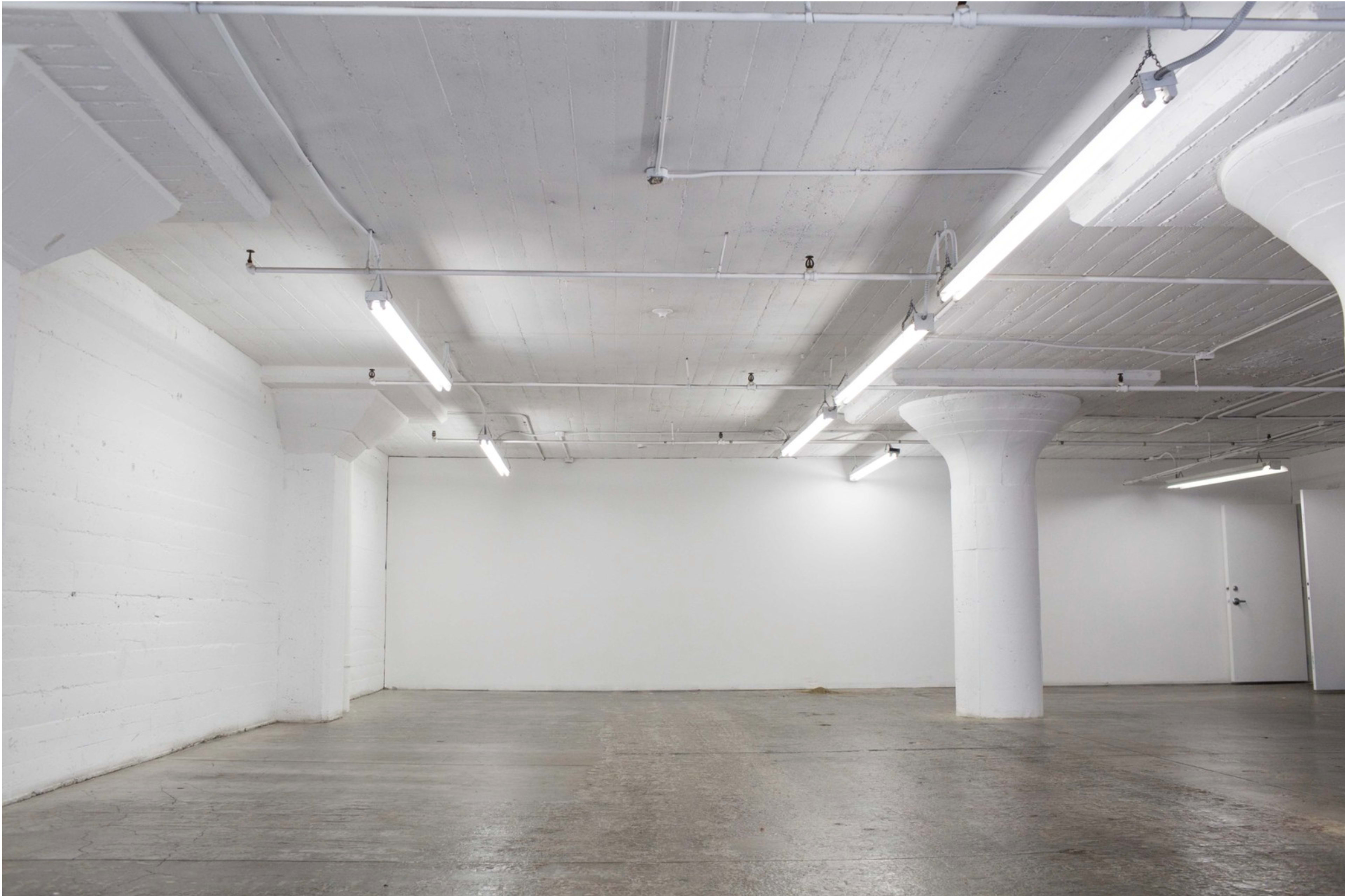 An empty industrial space features exposed concrete walls, columns, and fluorescent lighting.