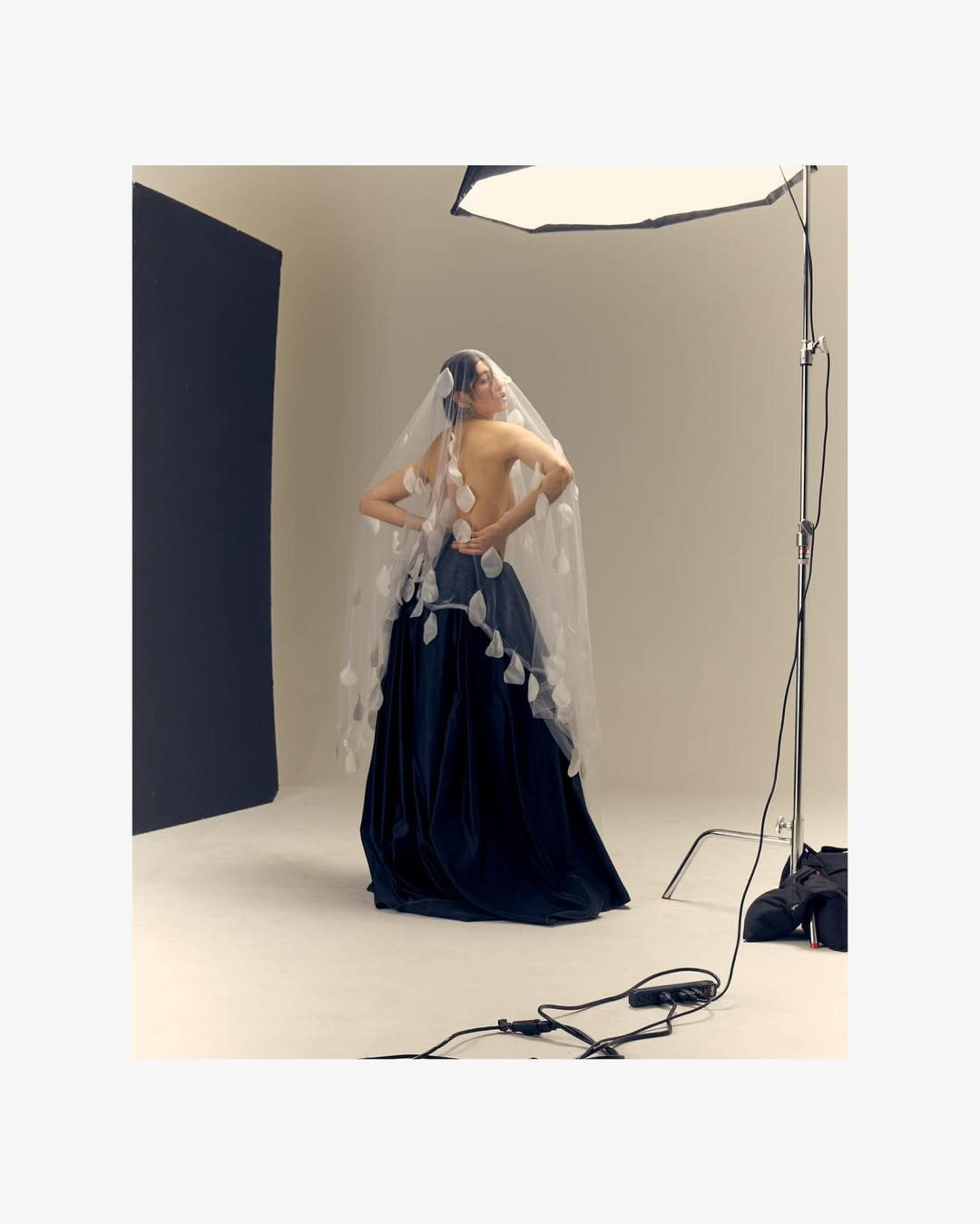 A model poses in a studio wearing a dark gown and a long veil adorned with circular decorations.