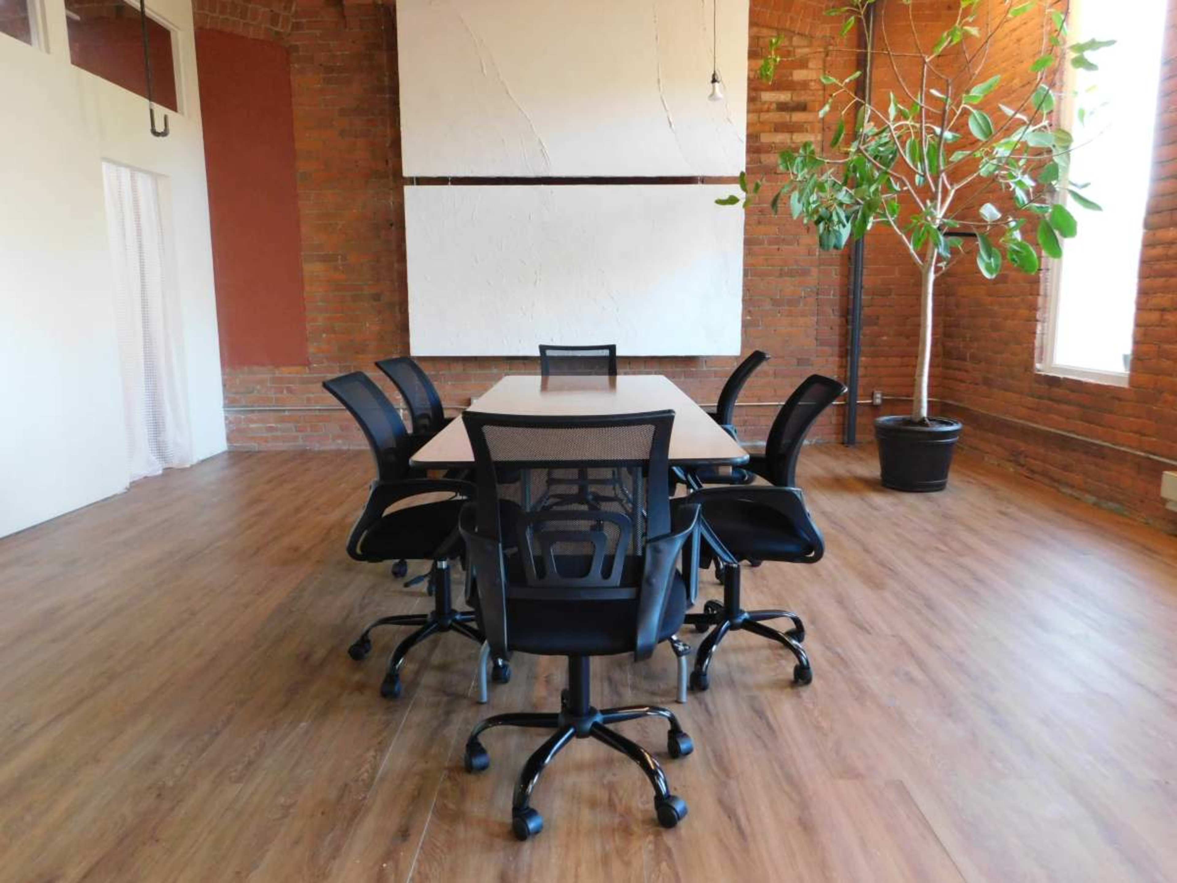 A conference room with a rectangular table surrounded by six black office chairs, set against a brick wall and featuring a large potted plant in one corner.