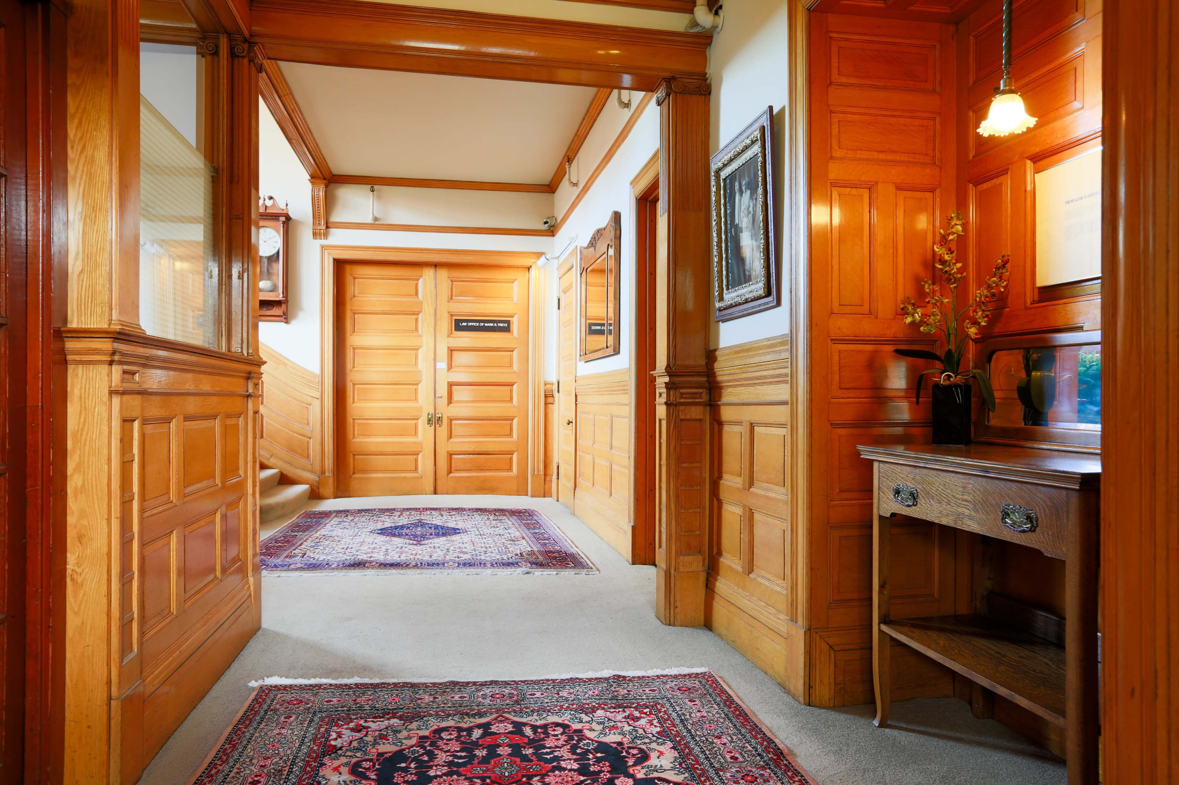 The image shows a hallway with wooden paneling, featuring a staircase, a decorative rug on the carpeted floor, and a small table against the wall.