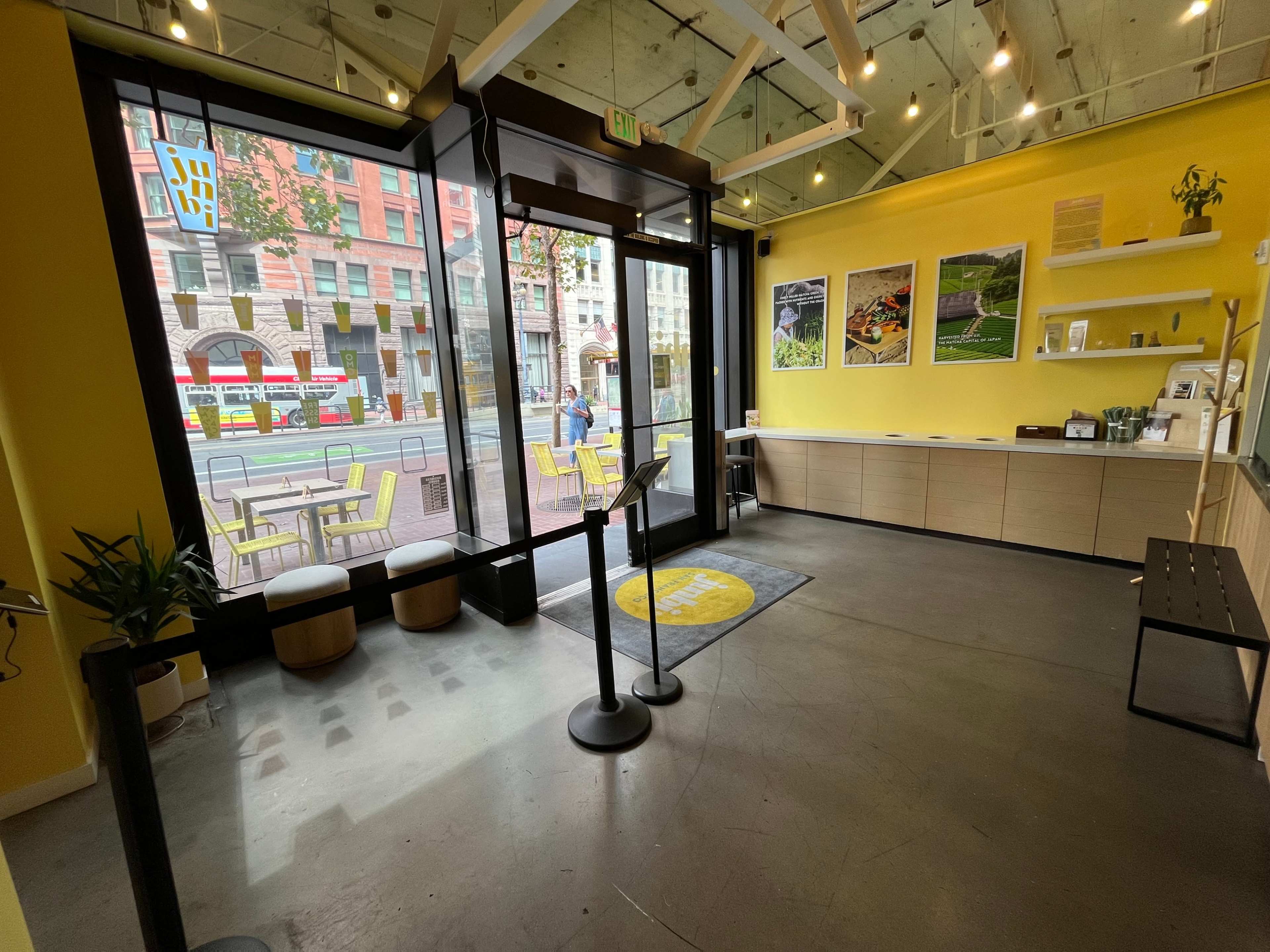 The image shows the interior of a brightly lit café with large windows facing an urban street, featuring a seating area and a service counter along one wall.