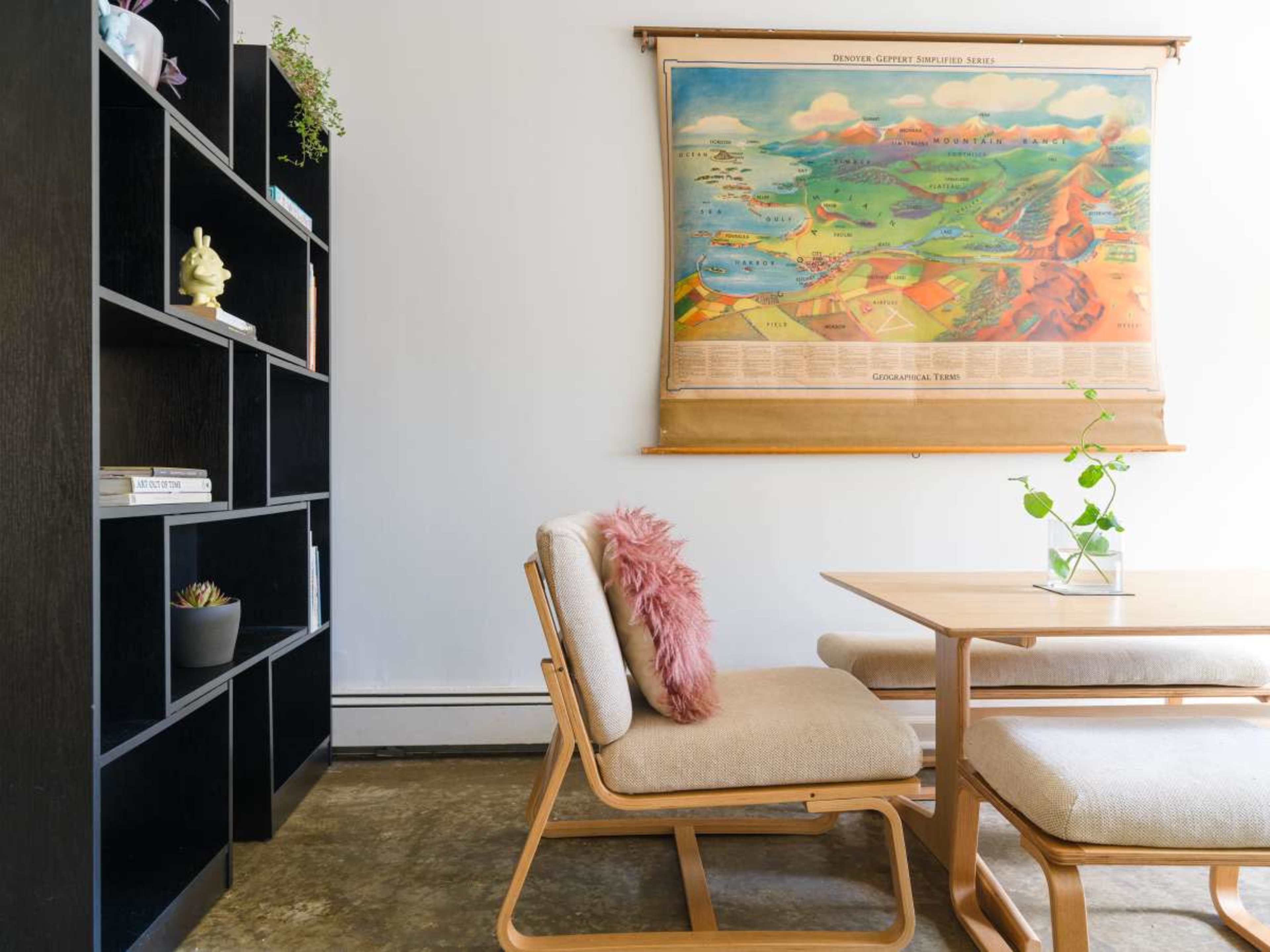A dining area features a wooden table and chairs, with a large vintage map hanging on the wall behind them and a black bookshelf to the left.