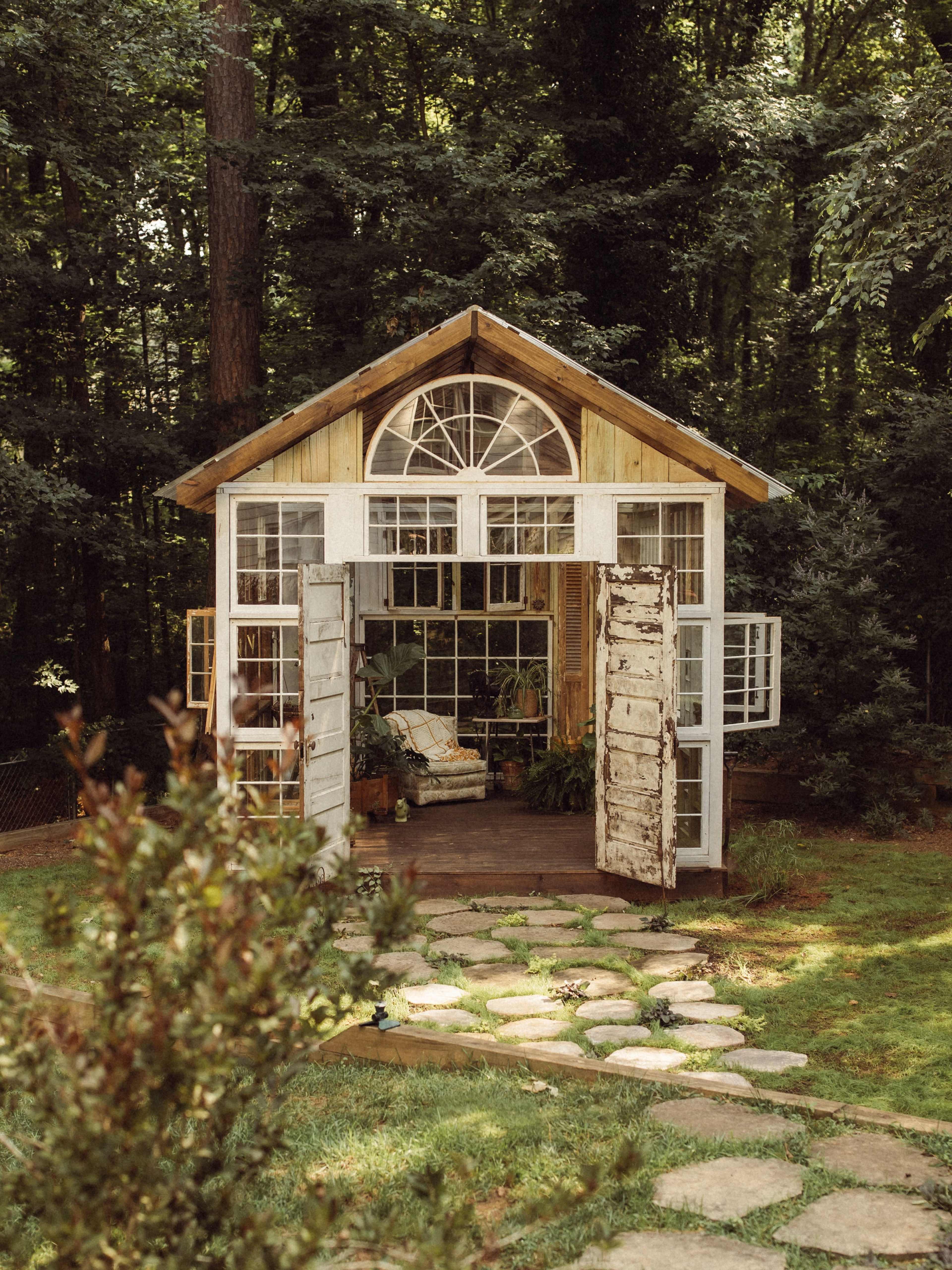 A small wooden shed with large windows and doors stands in a forested area, surrounded by greenery and stone pathways.
