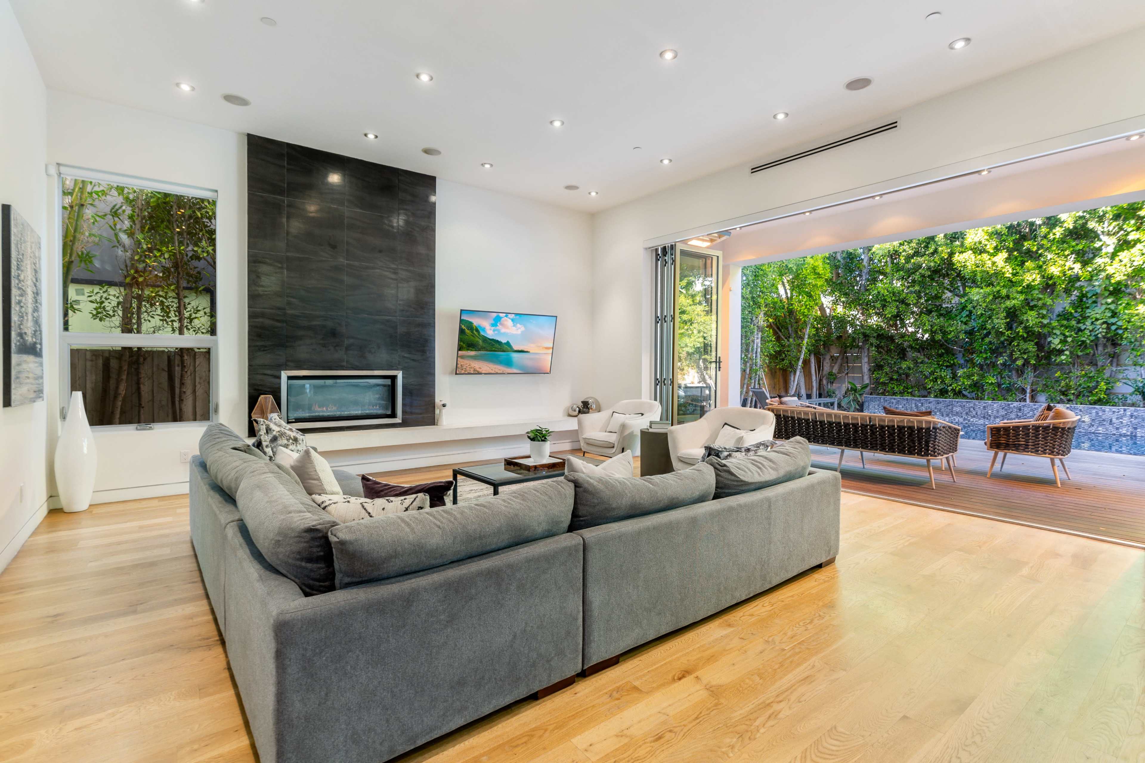 A modern living room with a spacious gray sofa, a sleek fireplace, and large sliding glass doors that open to an outdoor seating area surrounded by greenery.
