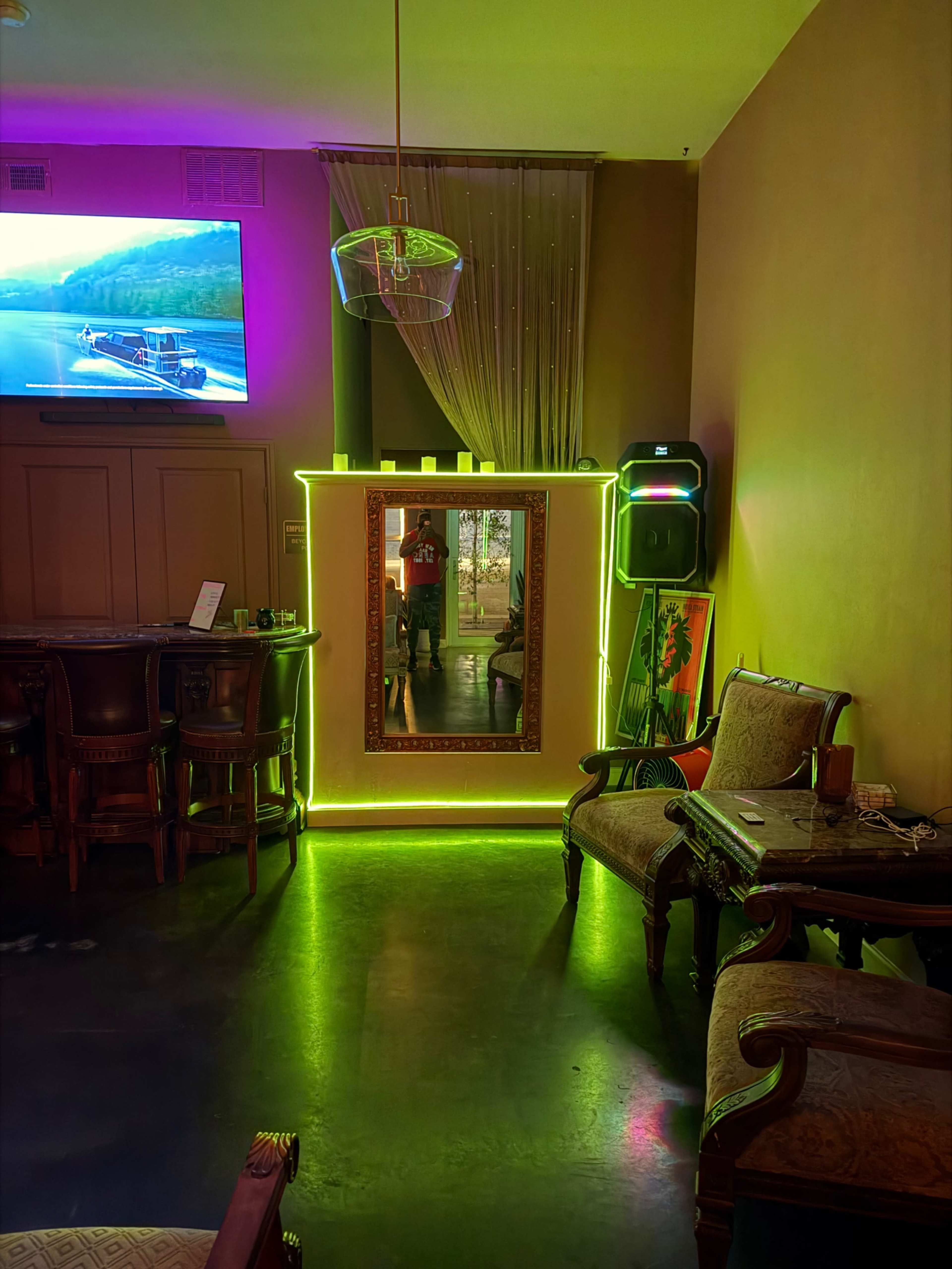 The image shows a cozy bar area featuring a brightly lit mirror with neon green accents, a television displaying a scenic view, and elegant seating arrangements.