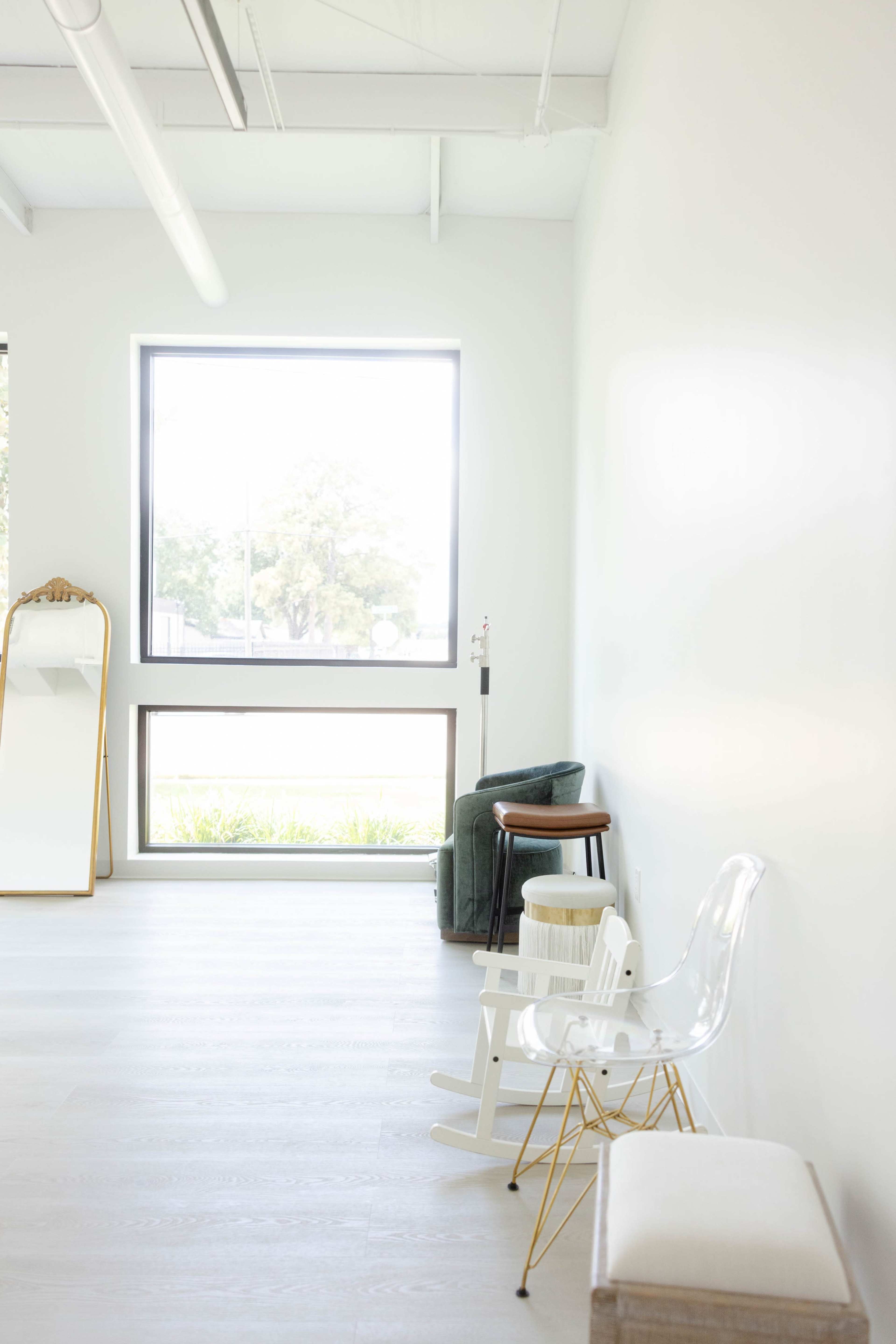 The image shows a bright, minimalistic room with a large window, a mirror, and various seating options including a rocking chair and stools.