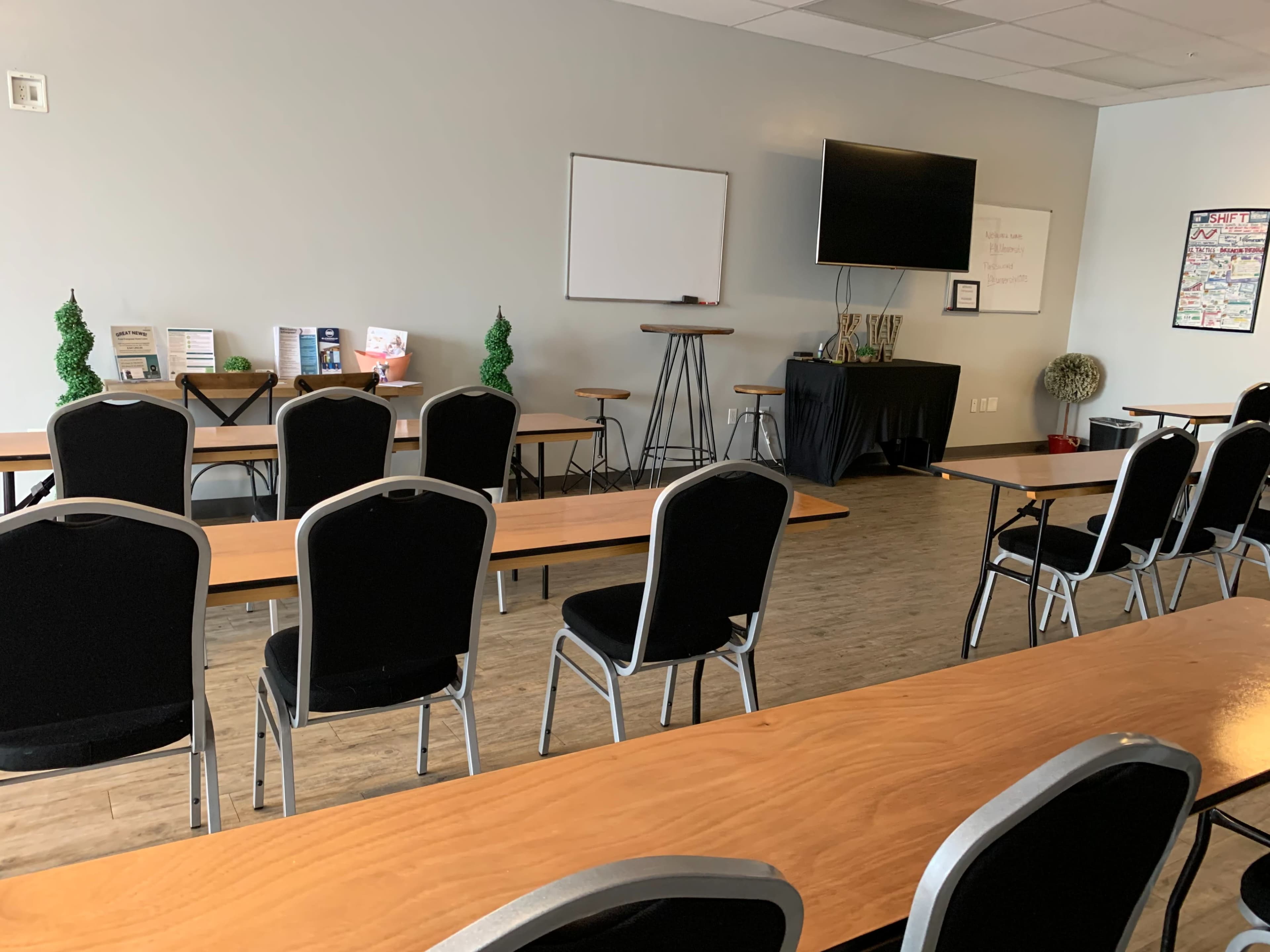 A seminar room with rows of black chairs facing a wooden table and a wall-mounted TV.