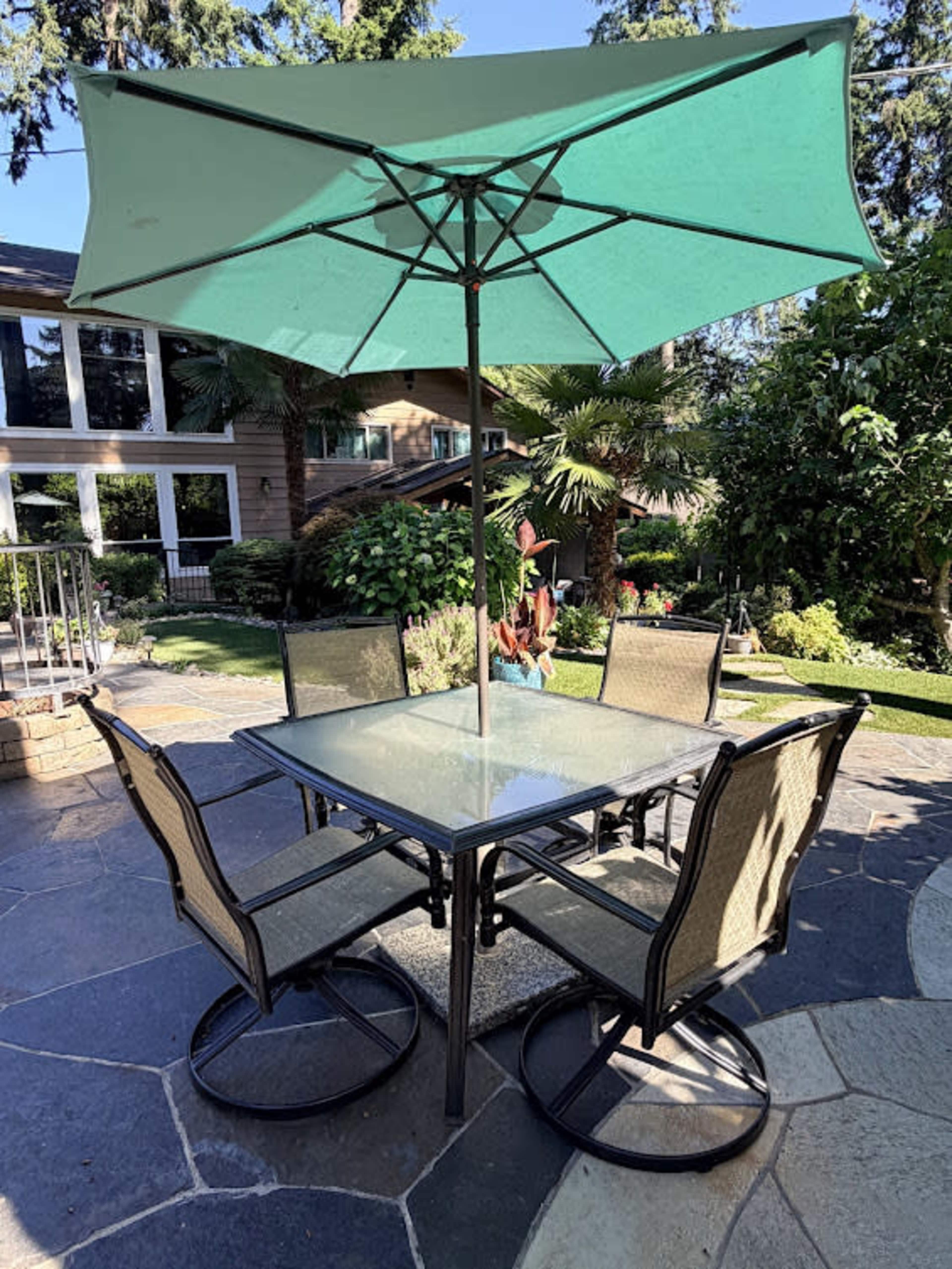 A glass-top dining table with four chairs and a green umbrella is set on a stone patio surrounded by lush greenery.