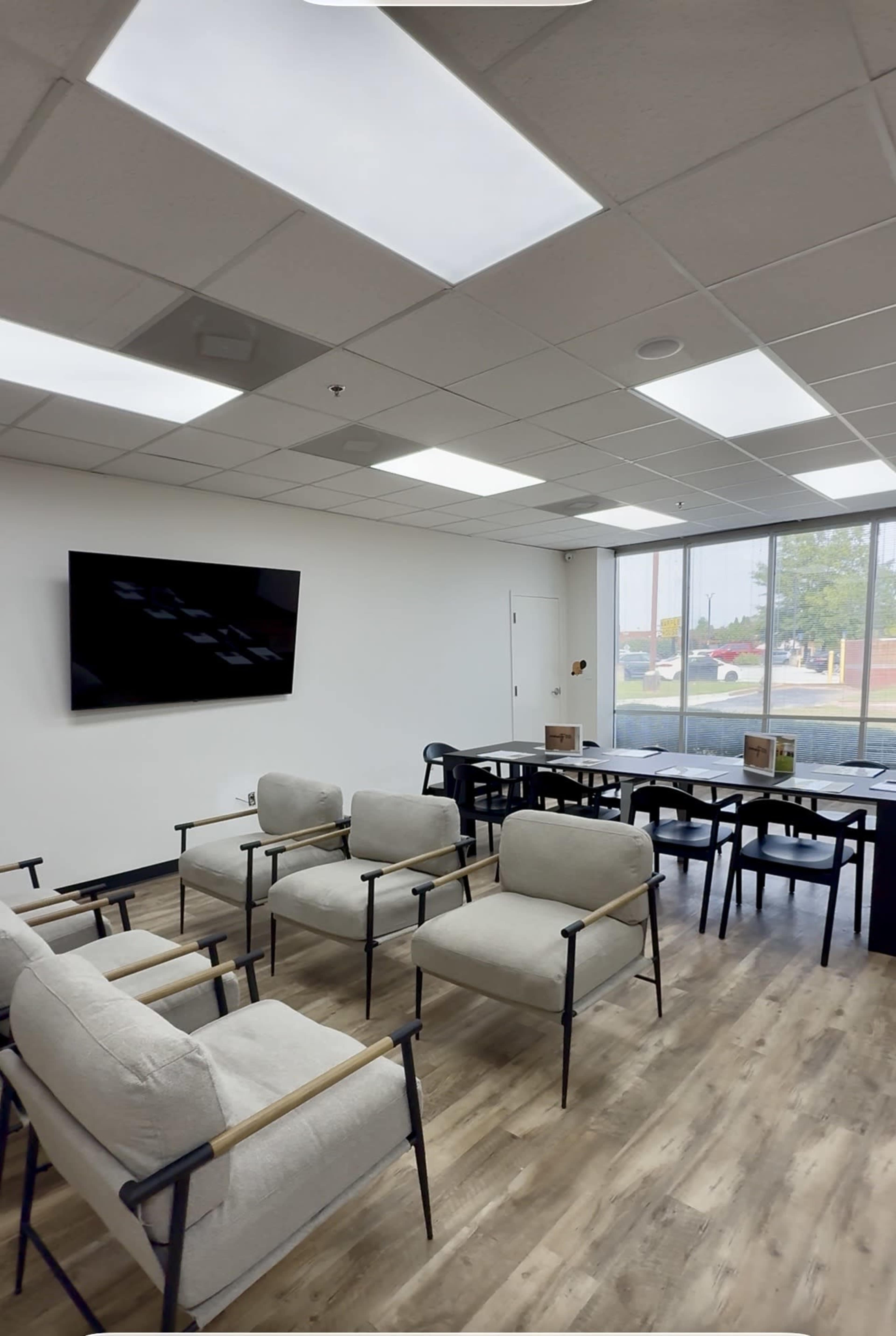 The image shows a modern waiting area with several light-colored chairs arranged around a small table, a wall-mounted television, and large windows allowing natural light to enter.