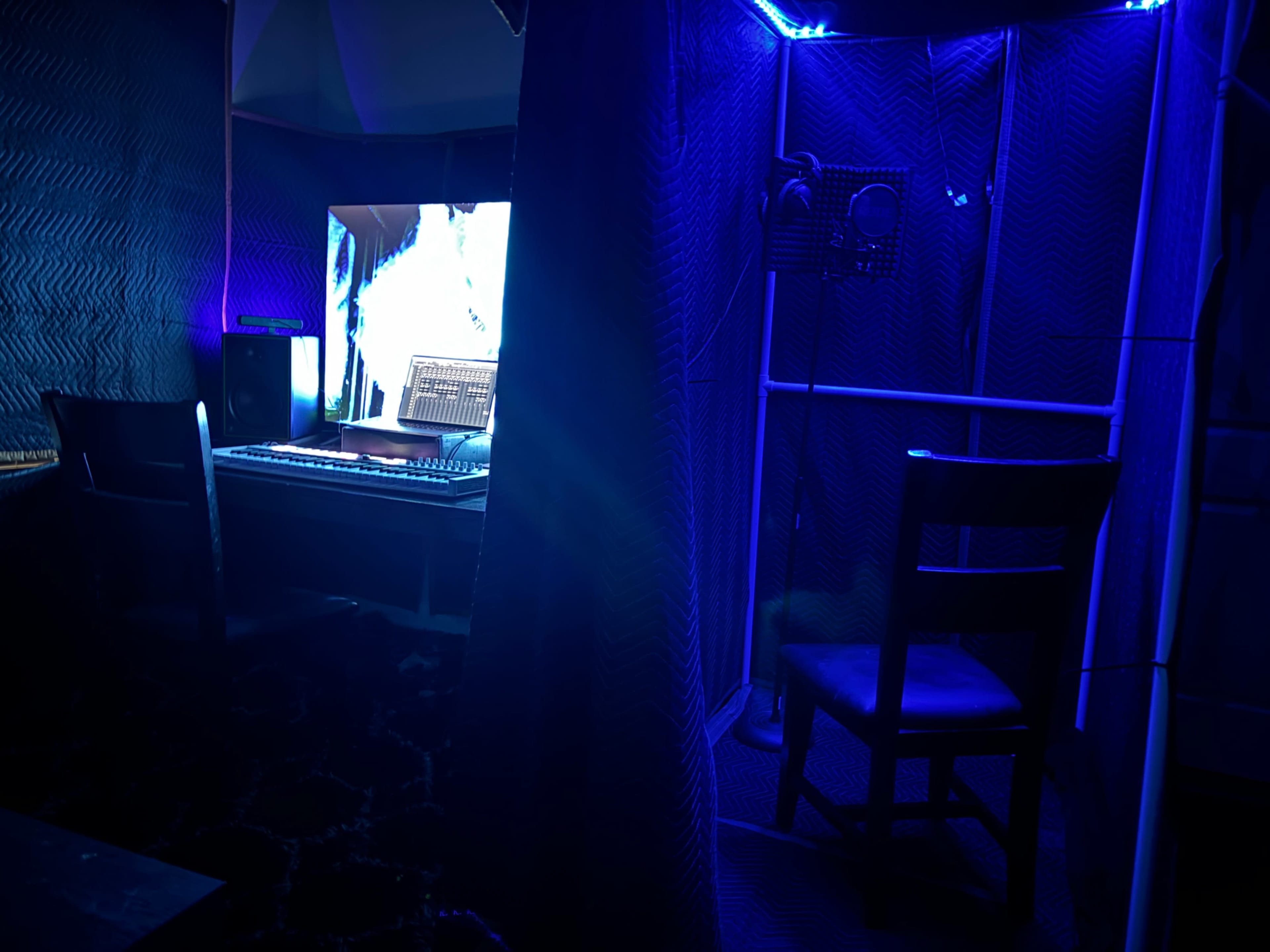 A desk with a computer and keyboard is set up within a soundproofed room illuminated by blue lights, with a chair positioned nearby.