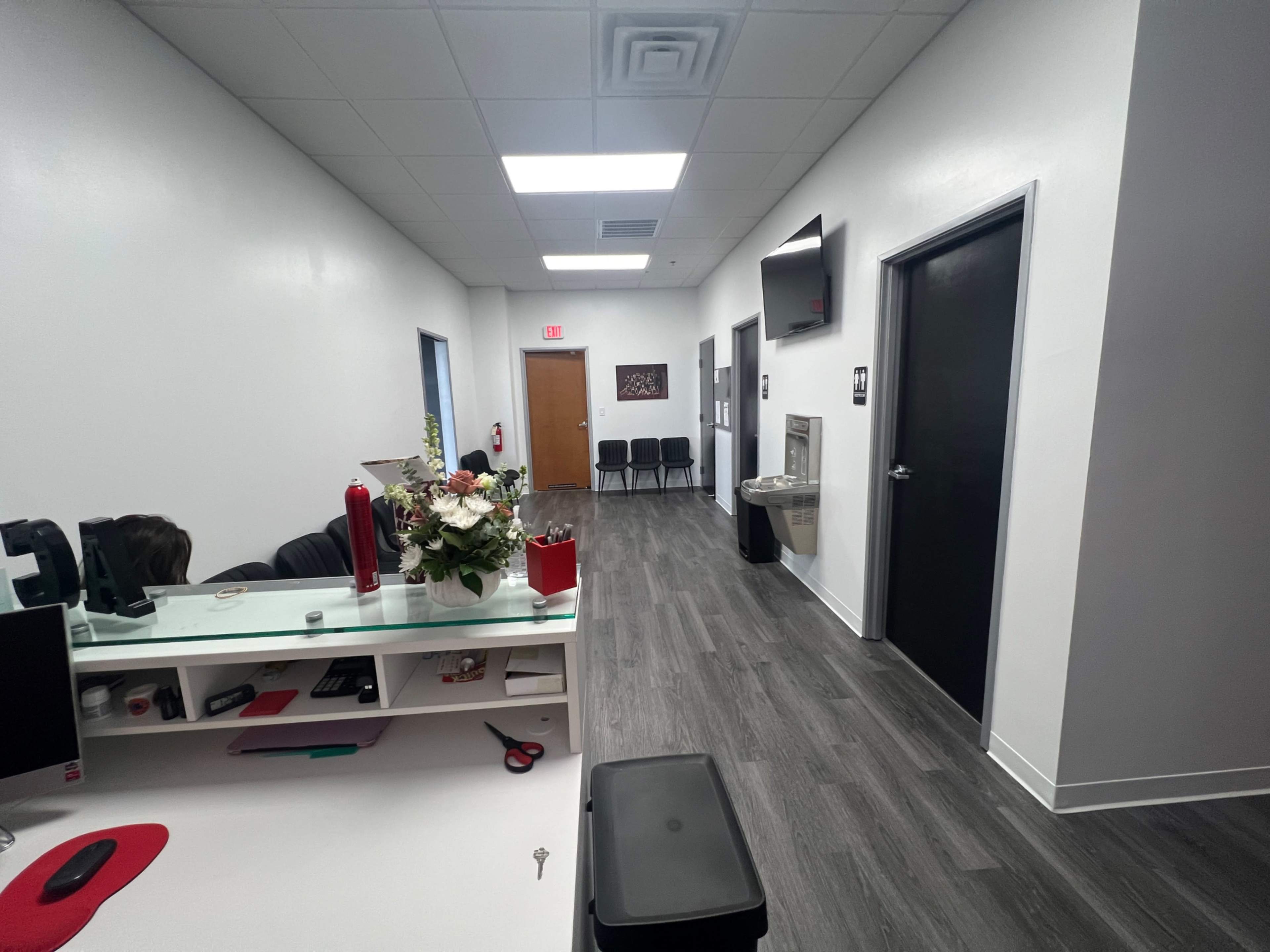 The image shows a modern office hallway with a reception area on the left, a water fountain on the right, and several chairs against the wall.