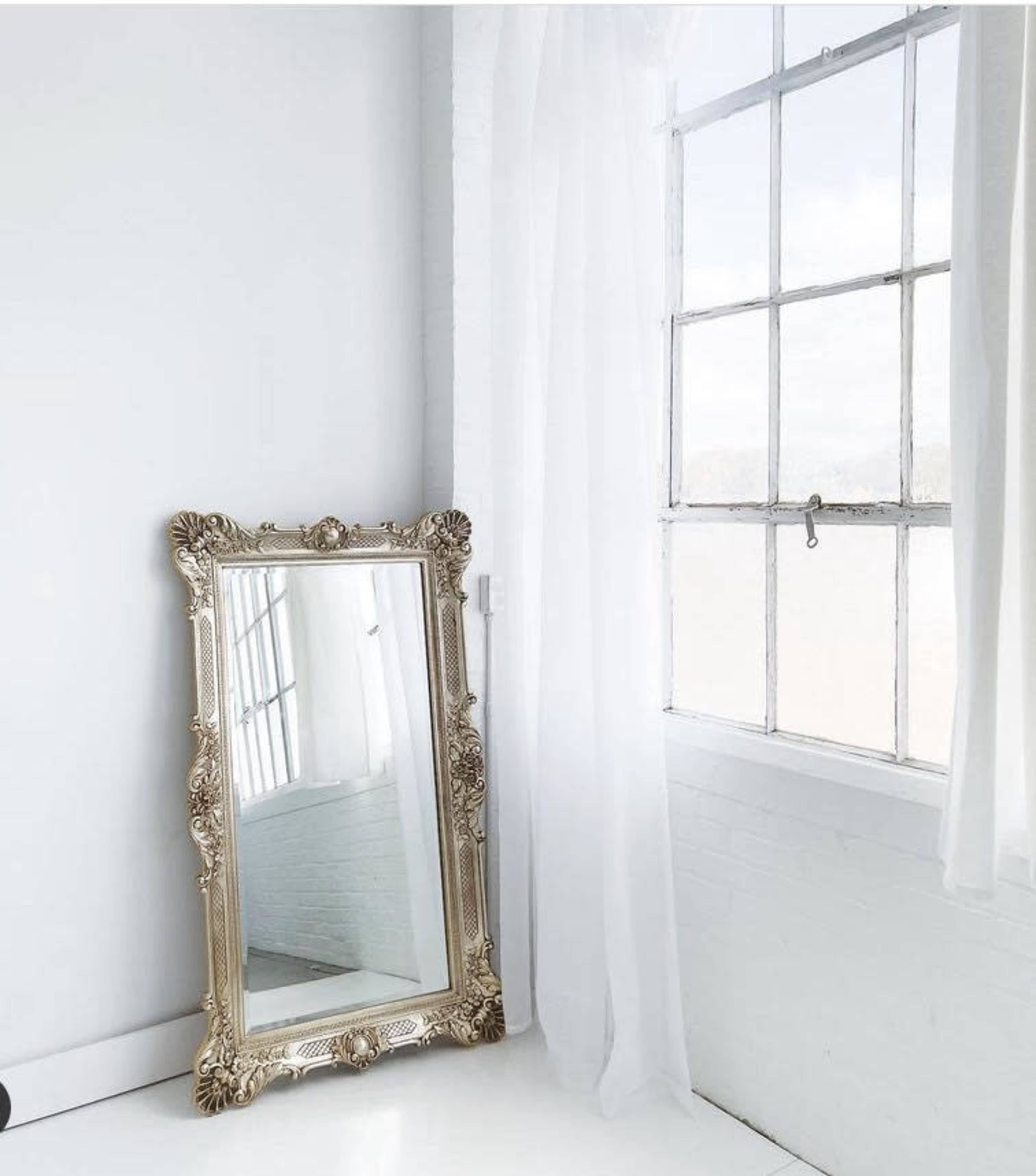 A large ornate gold-framed mirror stands next to a window with sheer white curtains, casting light in a bright, minimalistic room.