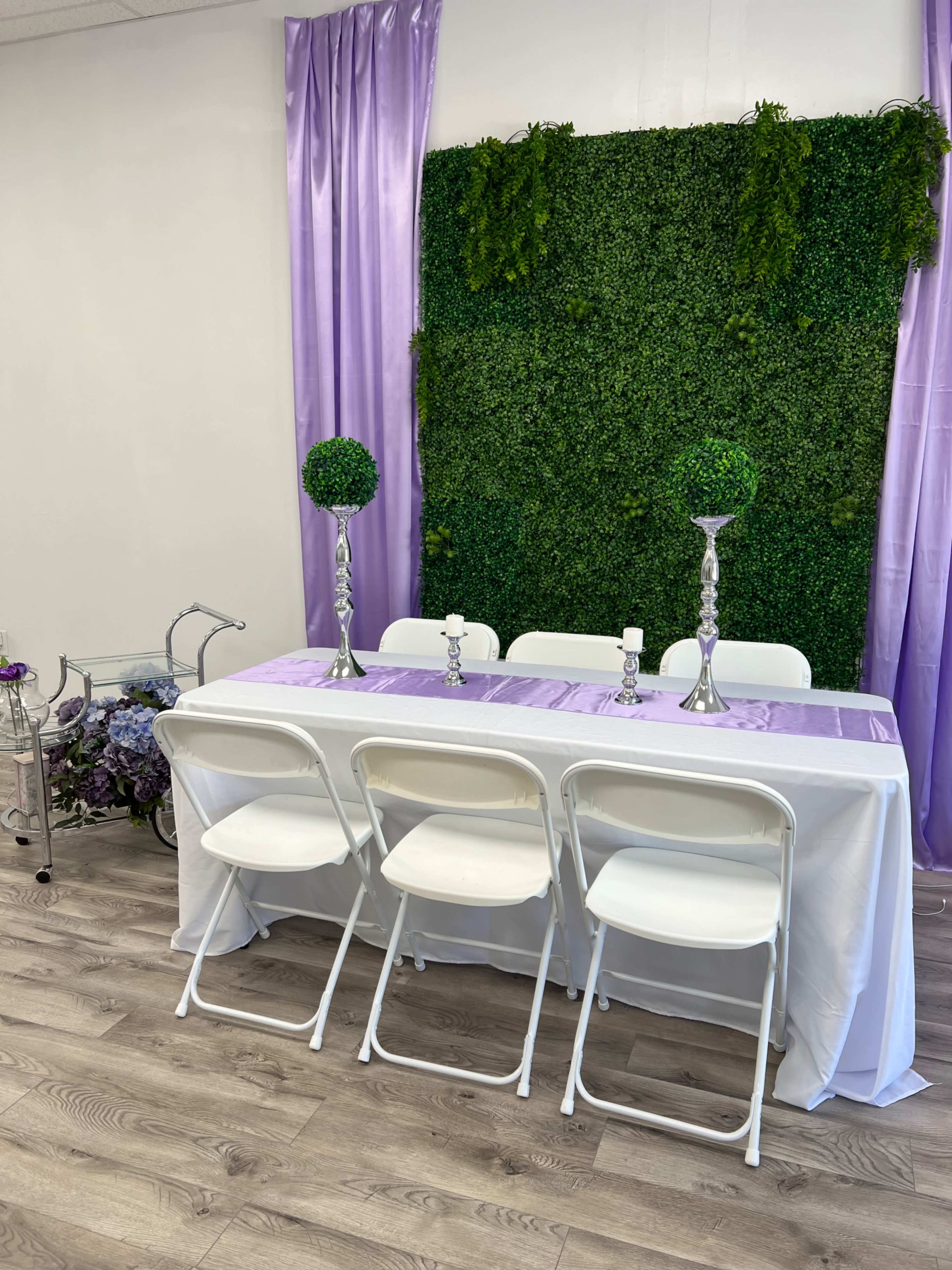 A decorated table with white chairs is set up in front of a green wall with decorative foliage, flanked by silver candle holders and topiary plants.