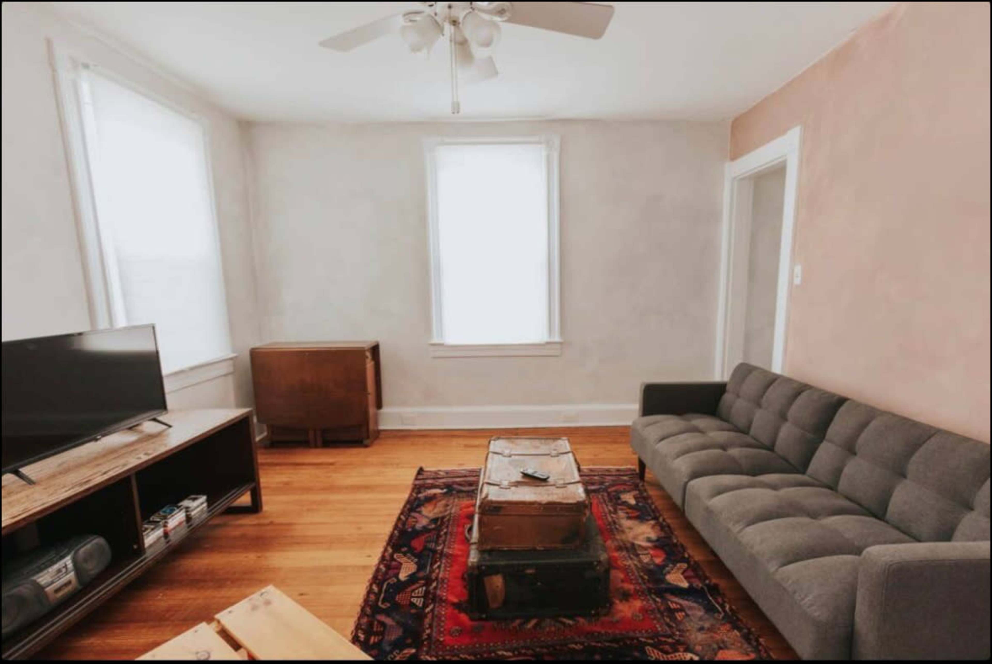 The image shows a living room with a gray sofa, a wooden coffee table with storage, a TV on a wooden stand, and two windows allowing natural light.