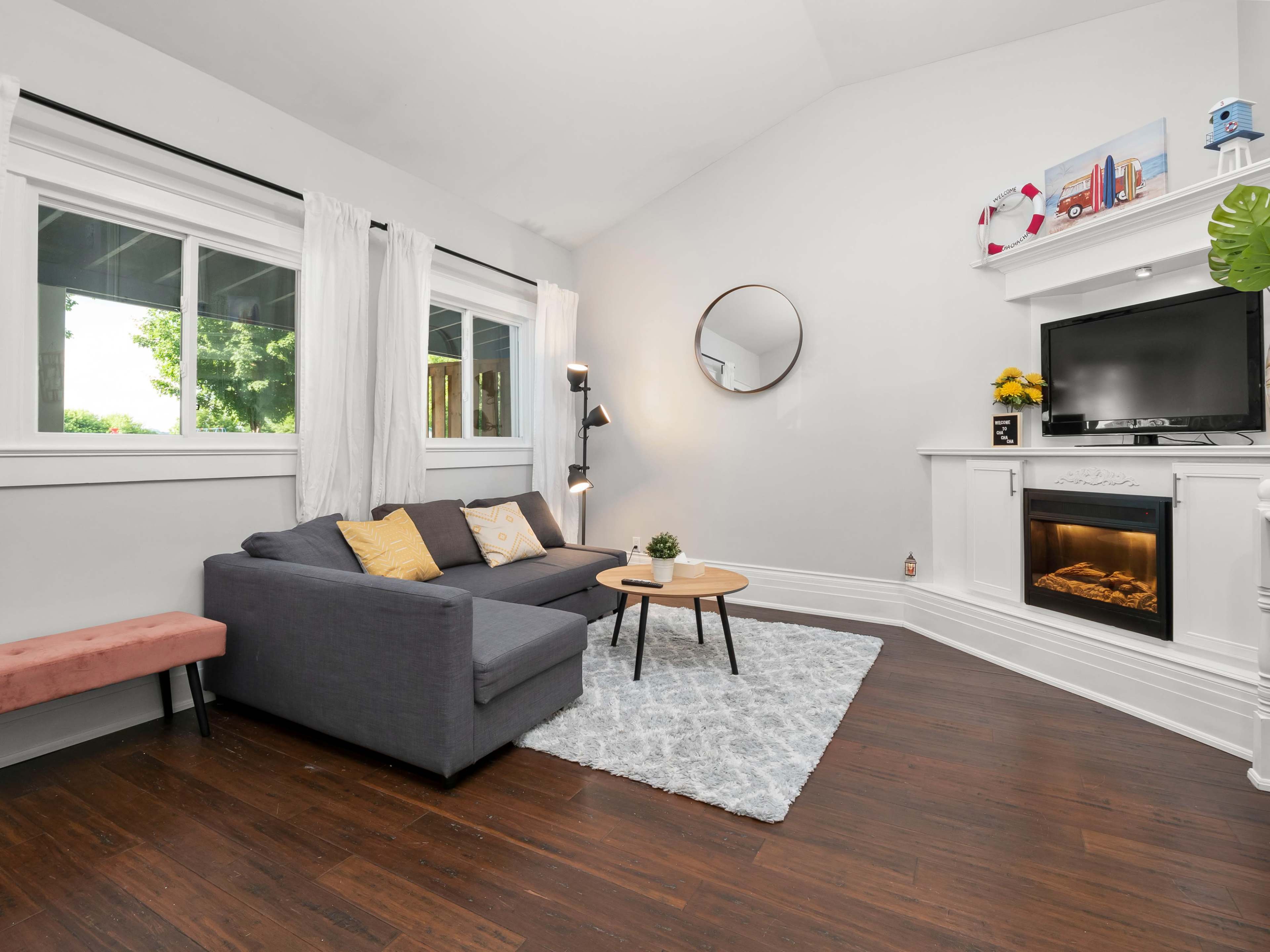 A modern living room with a gray couch, a round mirror, a television mounted on a white fireplace, and large windows allowing natural light to enter.