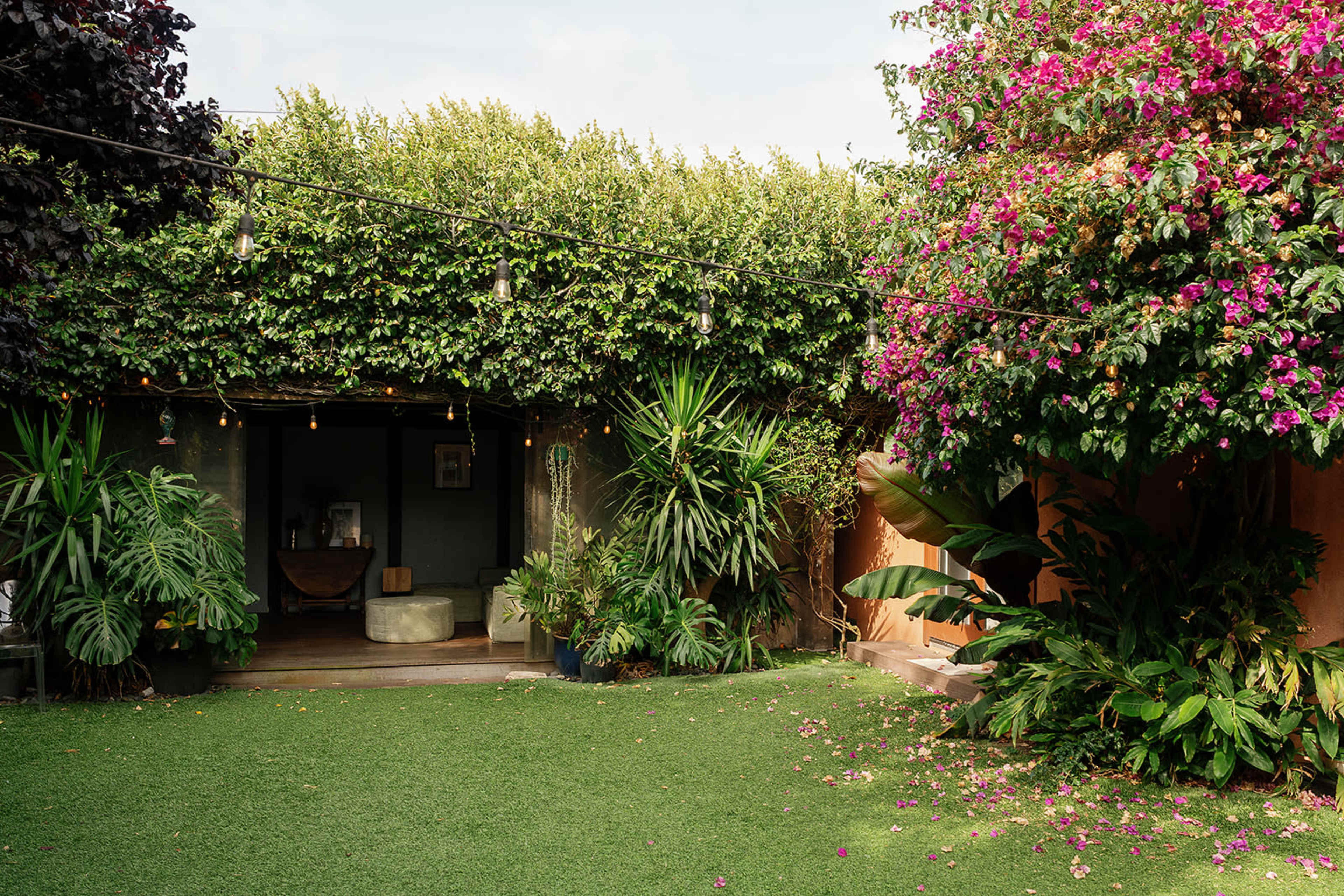 A lush garden features vibrant plants and flowers, surrounded by a green lawn and a shaded seating area with string lights.