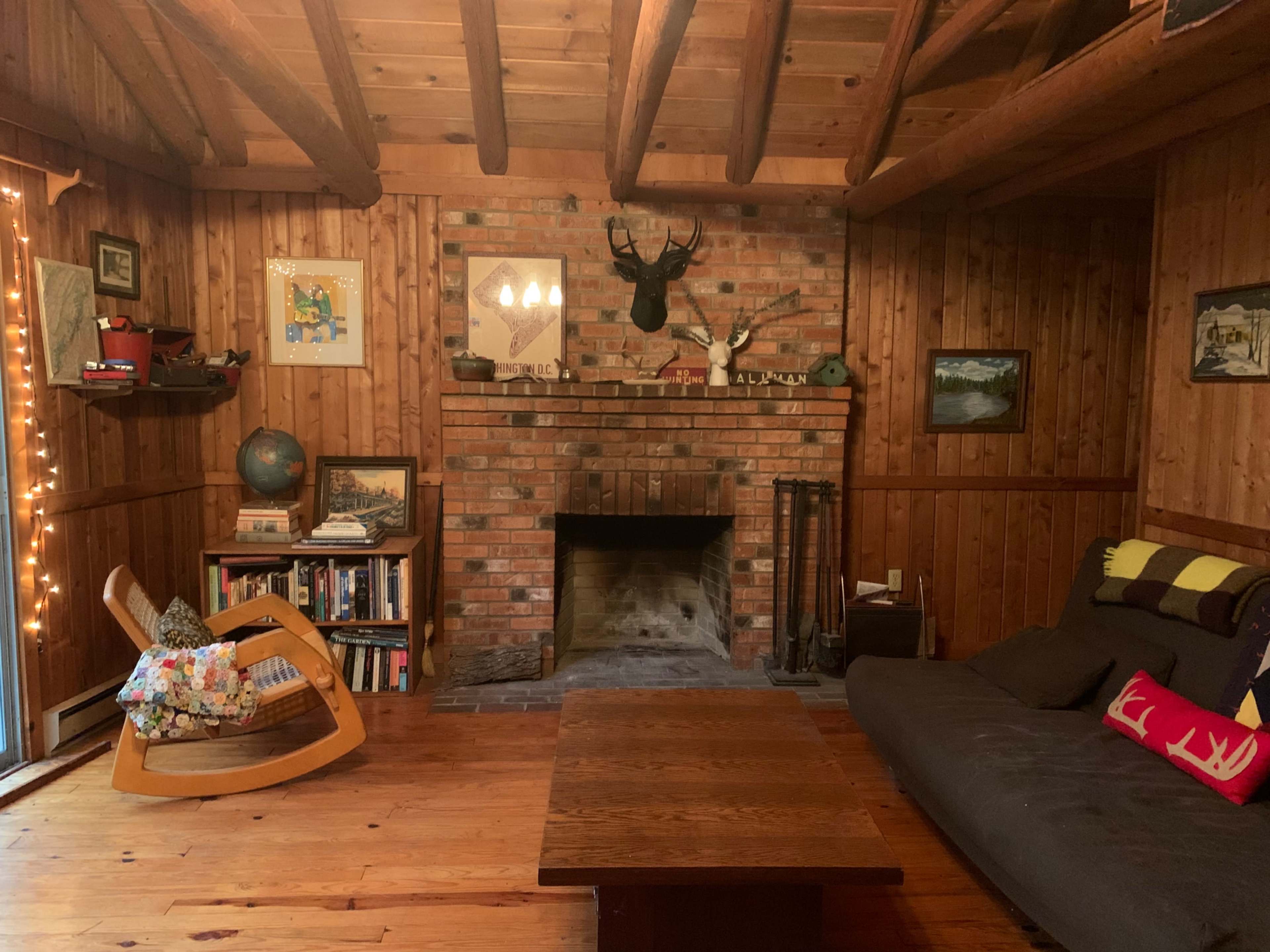 The living room features a brick fireplace, wooden beams, a rocking chair, and a wooden table surrounded by bookshelves and wall art.