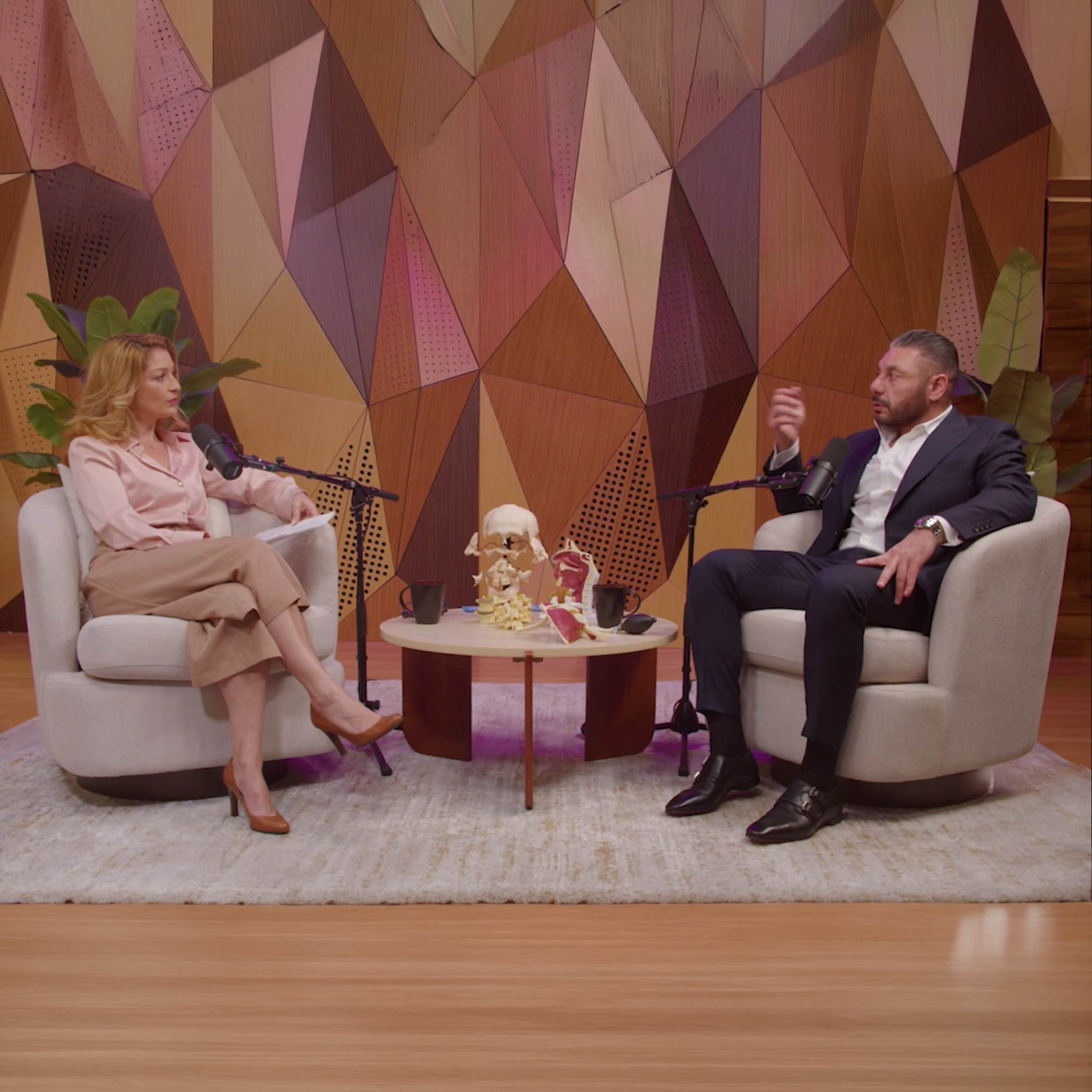 Two people are seated in modern armchairs in a stylish studio, engaging in conversation with microphones in front of them and a decorative table with various items nearby.