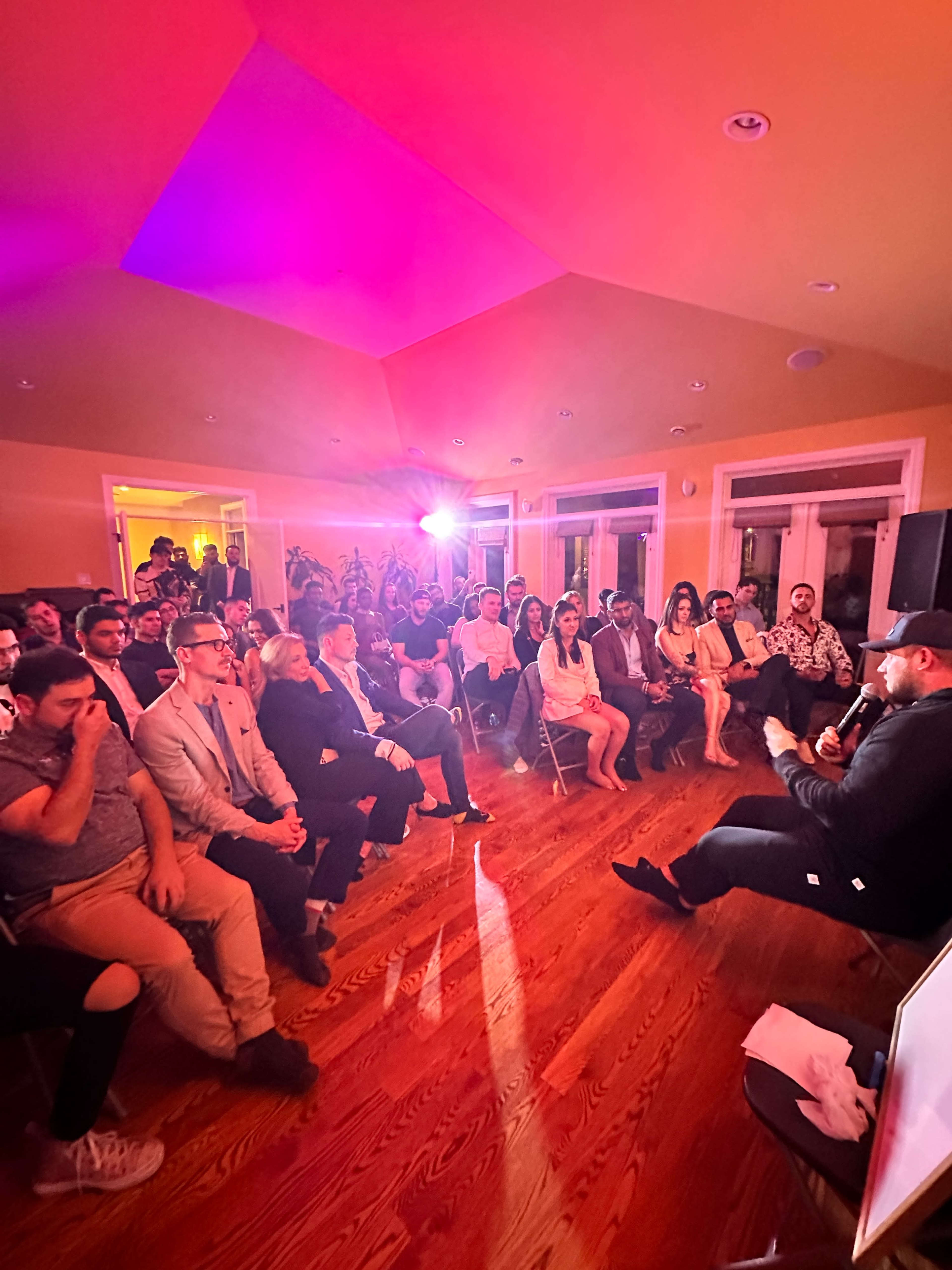 A crowd of seated people listens attentively to a speaker in a well-lit room with colorful lighting effects.