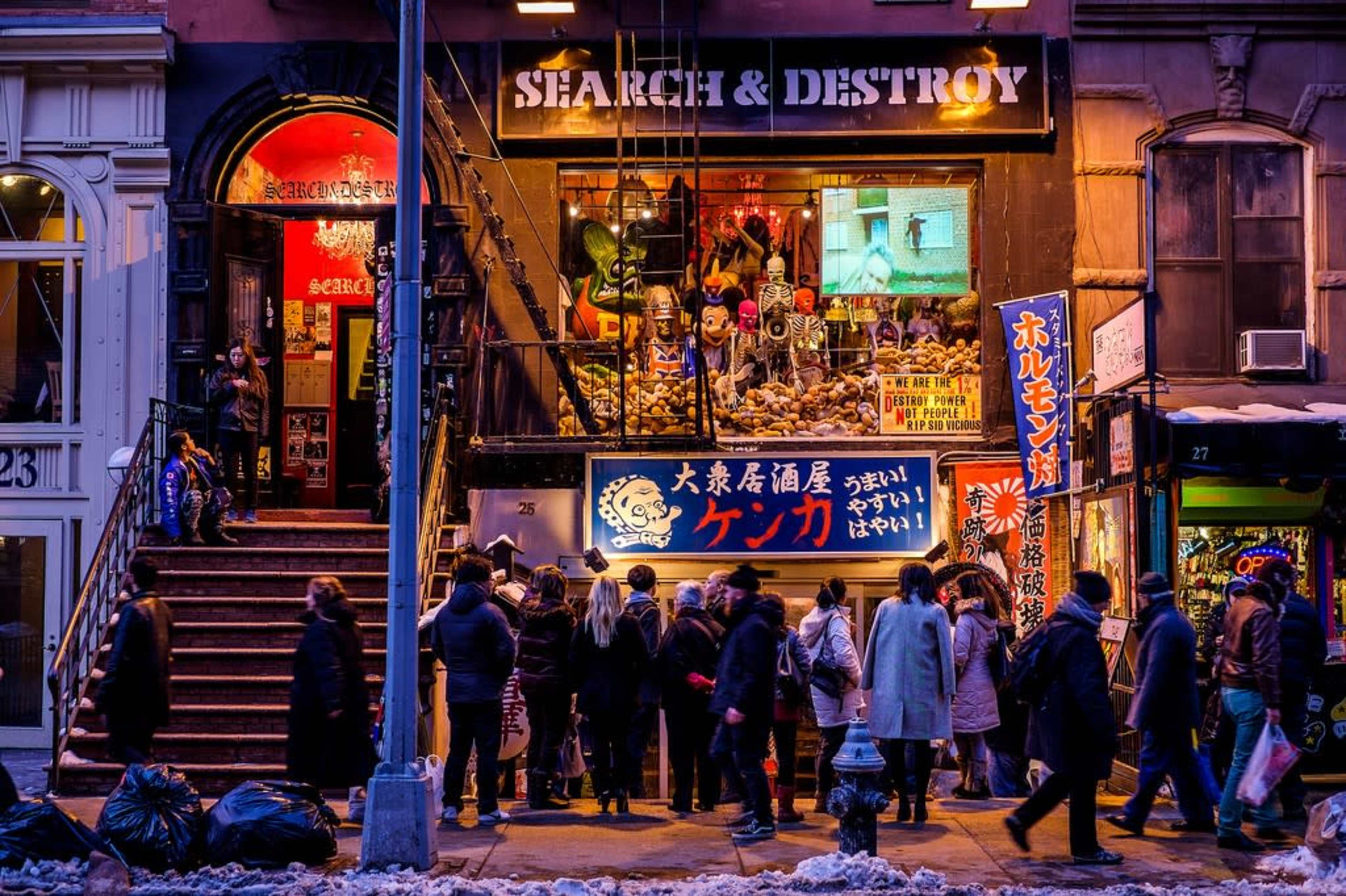 A crowd gathers outside a vibrant storefront named "SEARCH & DESTROY," adorned with colorful signs and window displays.