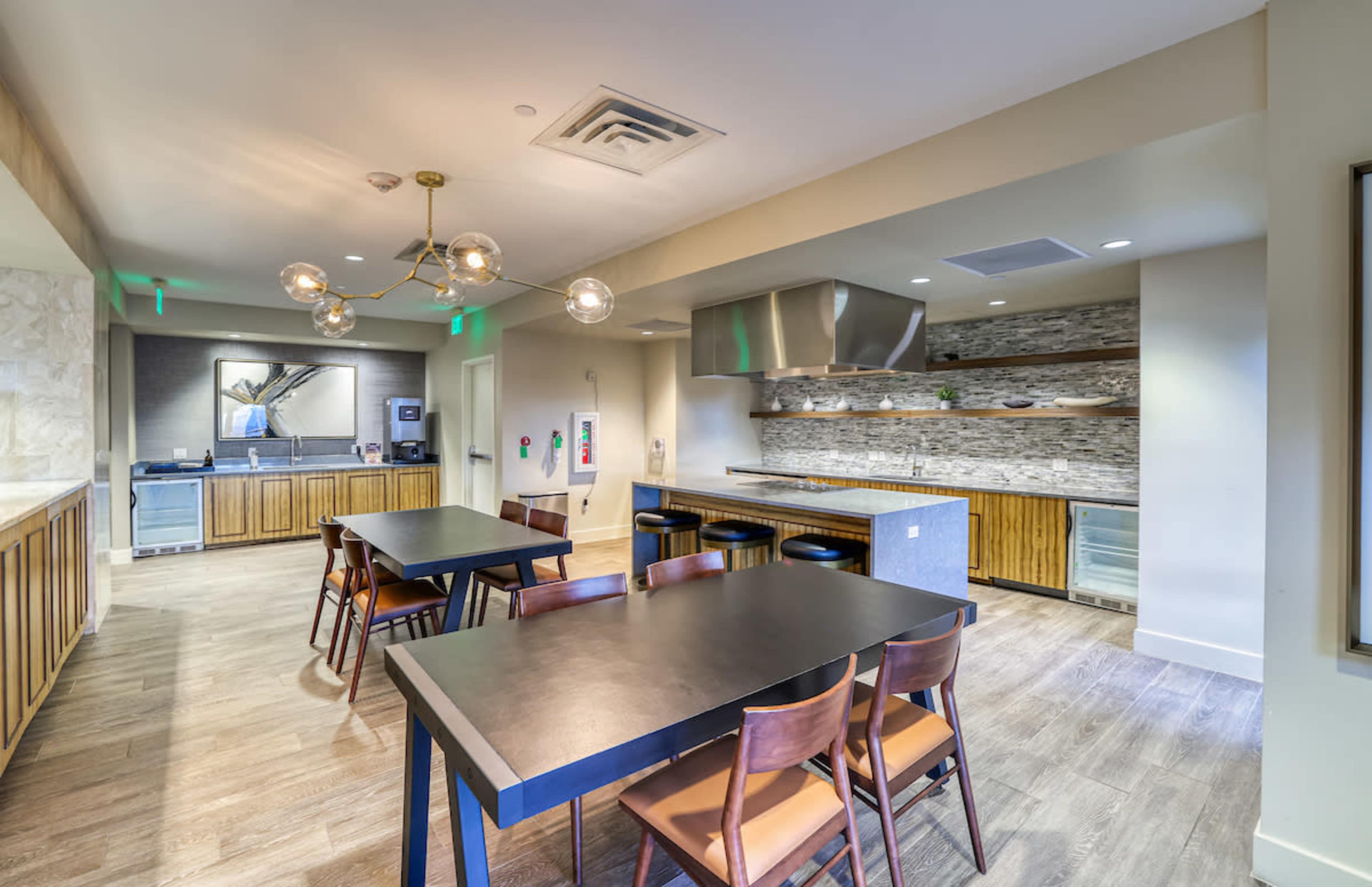 The image shows a modern kitchen and dining area with wooden cabinetry, a large table, and a stainless steel cooking range.