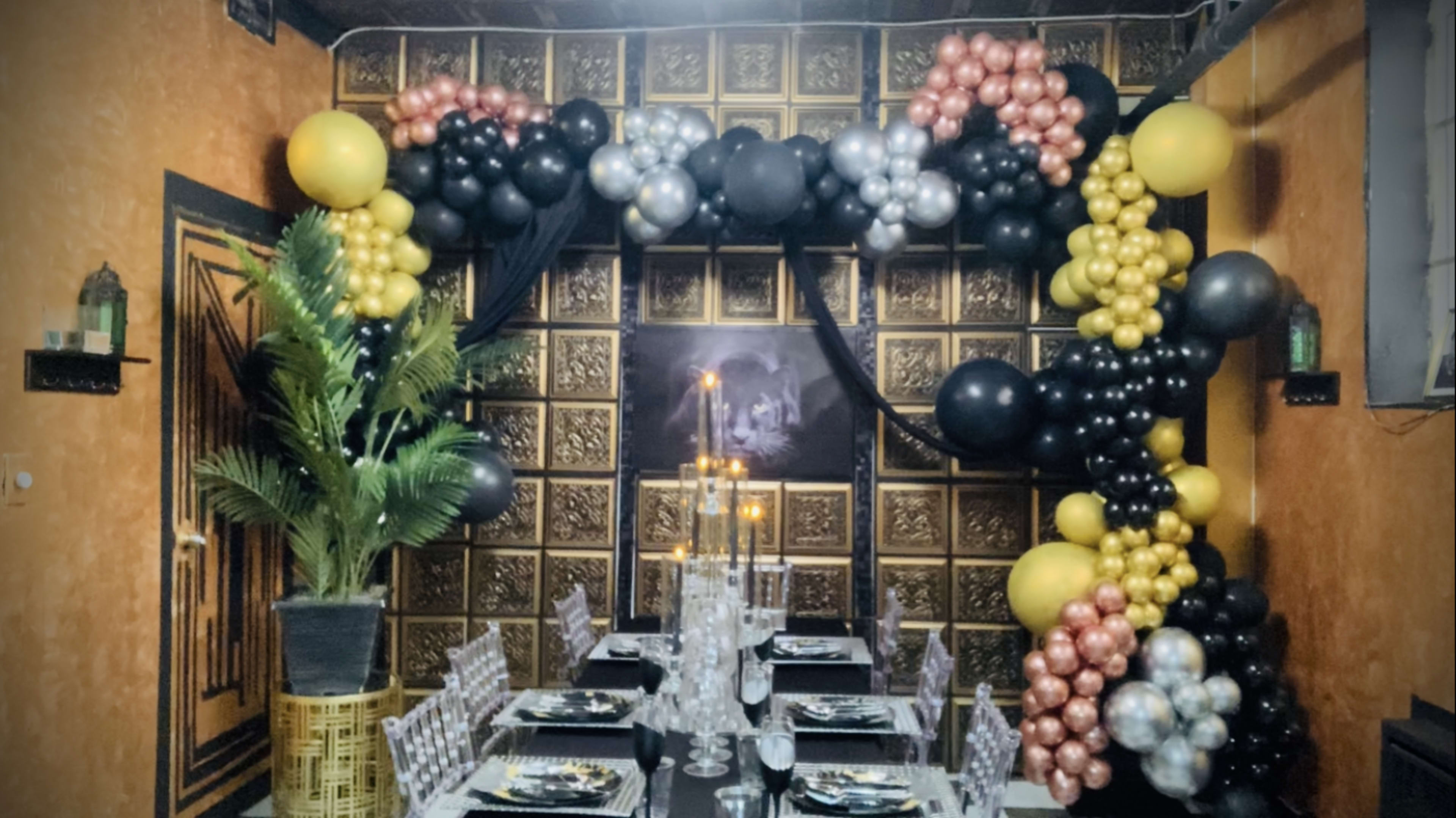 A decorated dining area featuring a balloon arch in black, gold, silver, and rose gold colors, with a table set for a formal meal underneath.