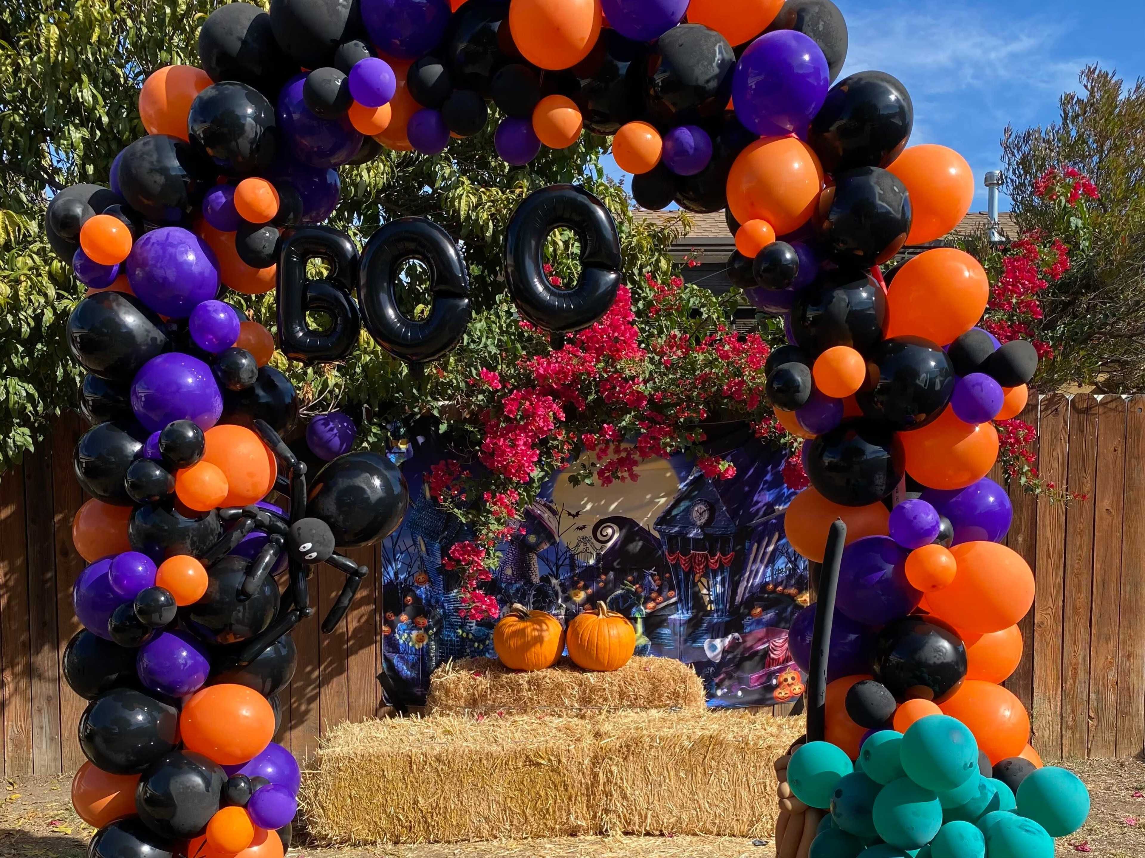 A decorative arch made of orange, black, and purple balloons frames a display of pumpkins on hay bales, set against a backdrop of flowers and Halloween-themed decorations.