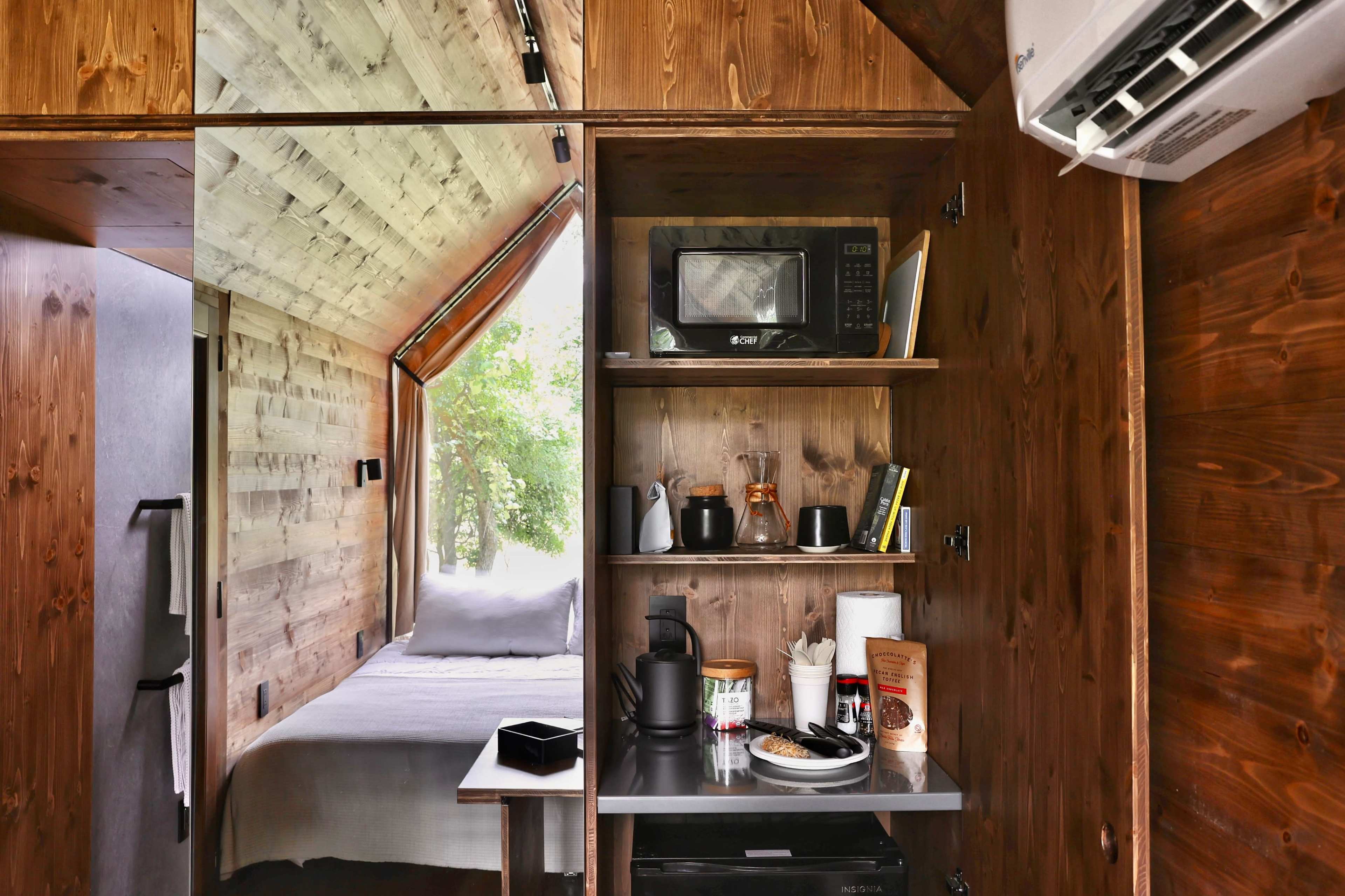 The image shows a compact wooden interior featuring a kitchenette with a microwave, coffee maker, and food items, alongside a small bedroom area with a bed.