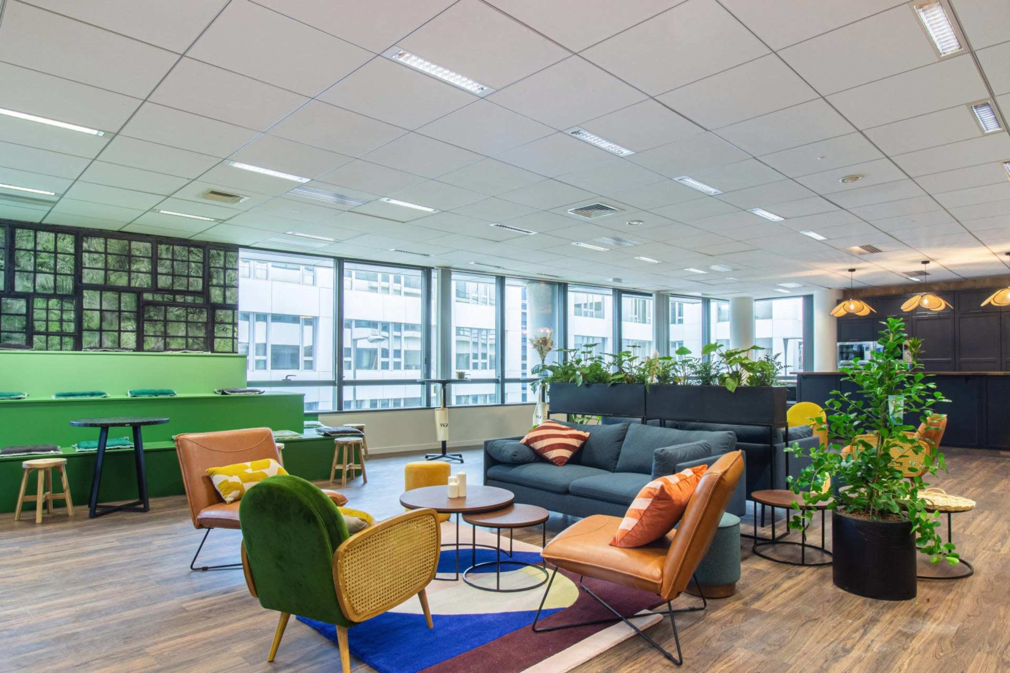 A modern office lounge area with comfortable seating, green plants, and large windows allowing natural light.