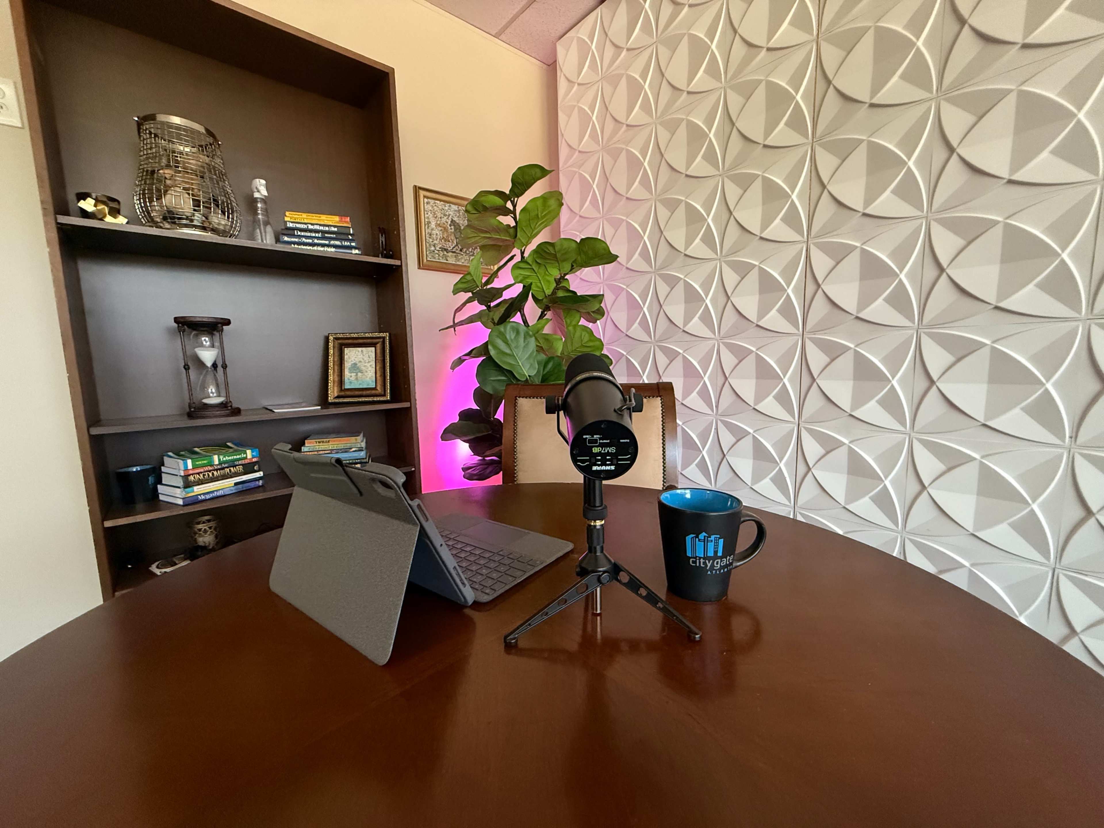 The image shows a small workspace featuring a microphone on a stand, a tablet in a case, a blue mug, and a bookshelf with books and decorative items against a textured wall.