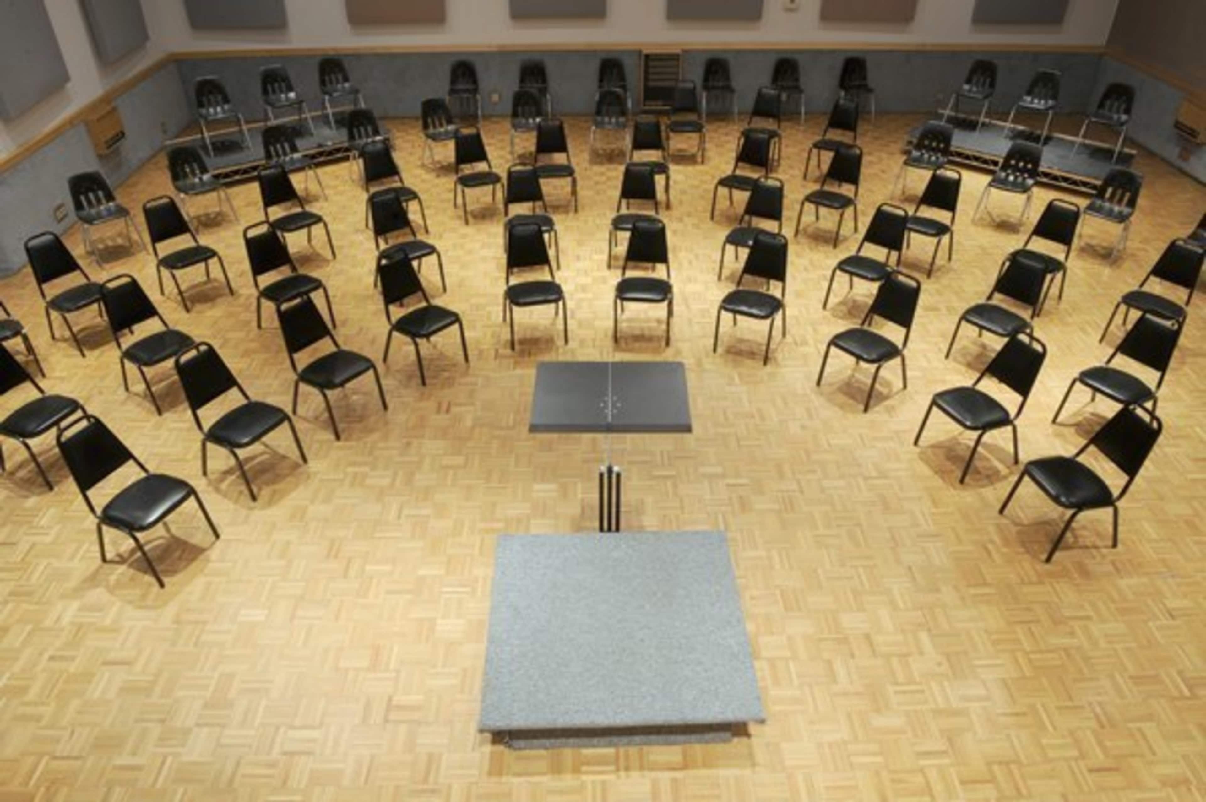 A circular arrangement of black chairs surrounds a central gray table on a wooden floor in a rehearsal space.