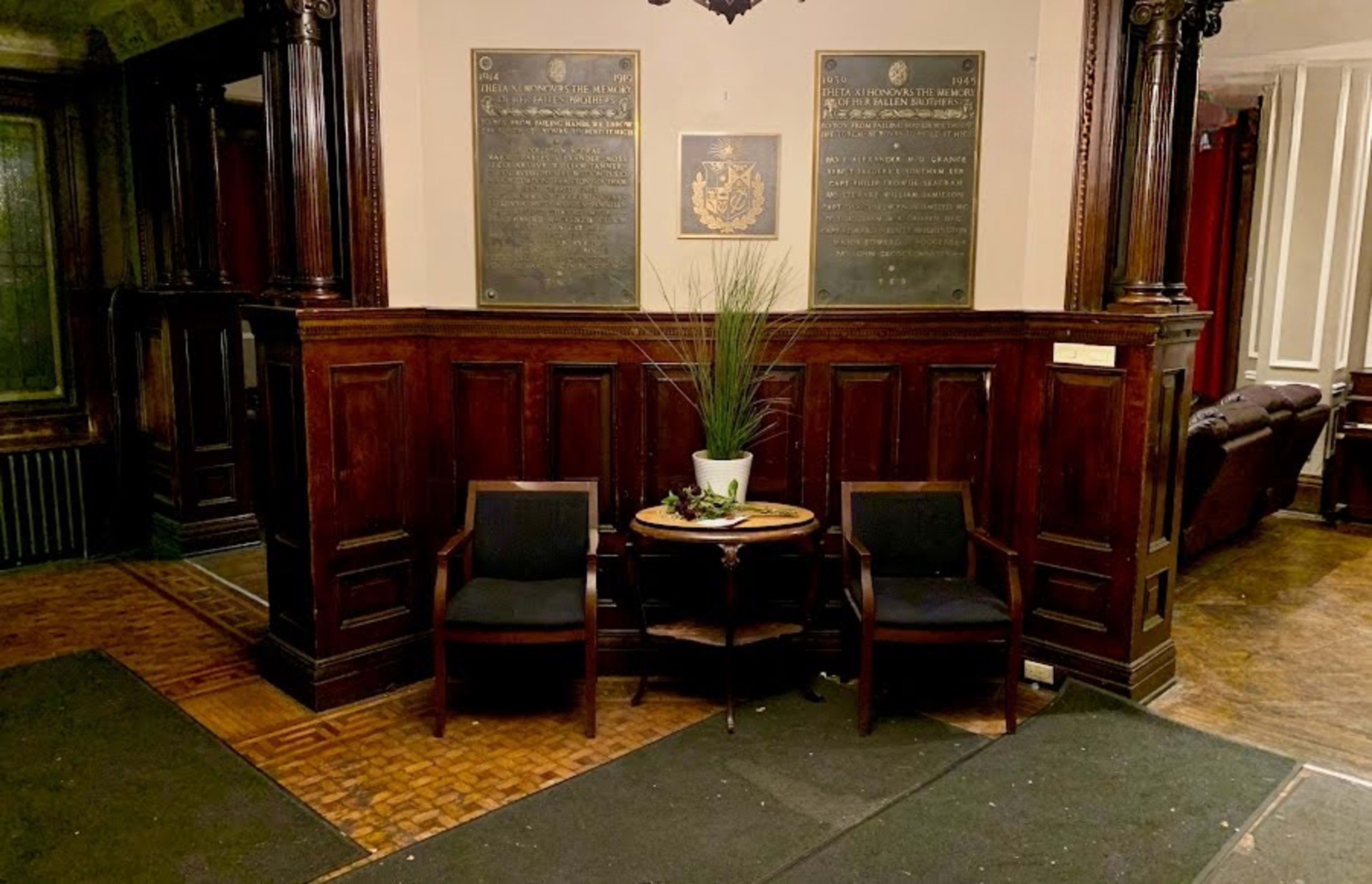 The image shows a wooden-paneled lobby with two chairs flanking a small round table, accompanied by potted plants and two plaques mounted on the wall.