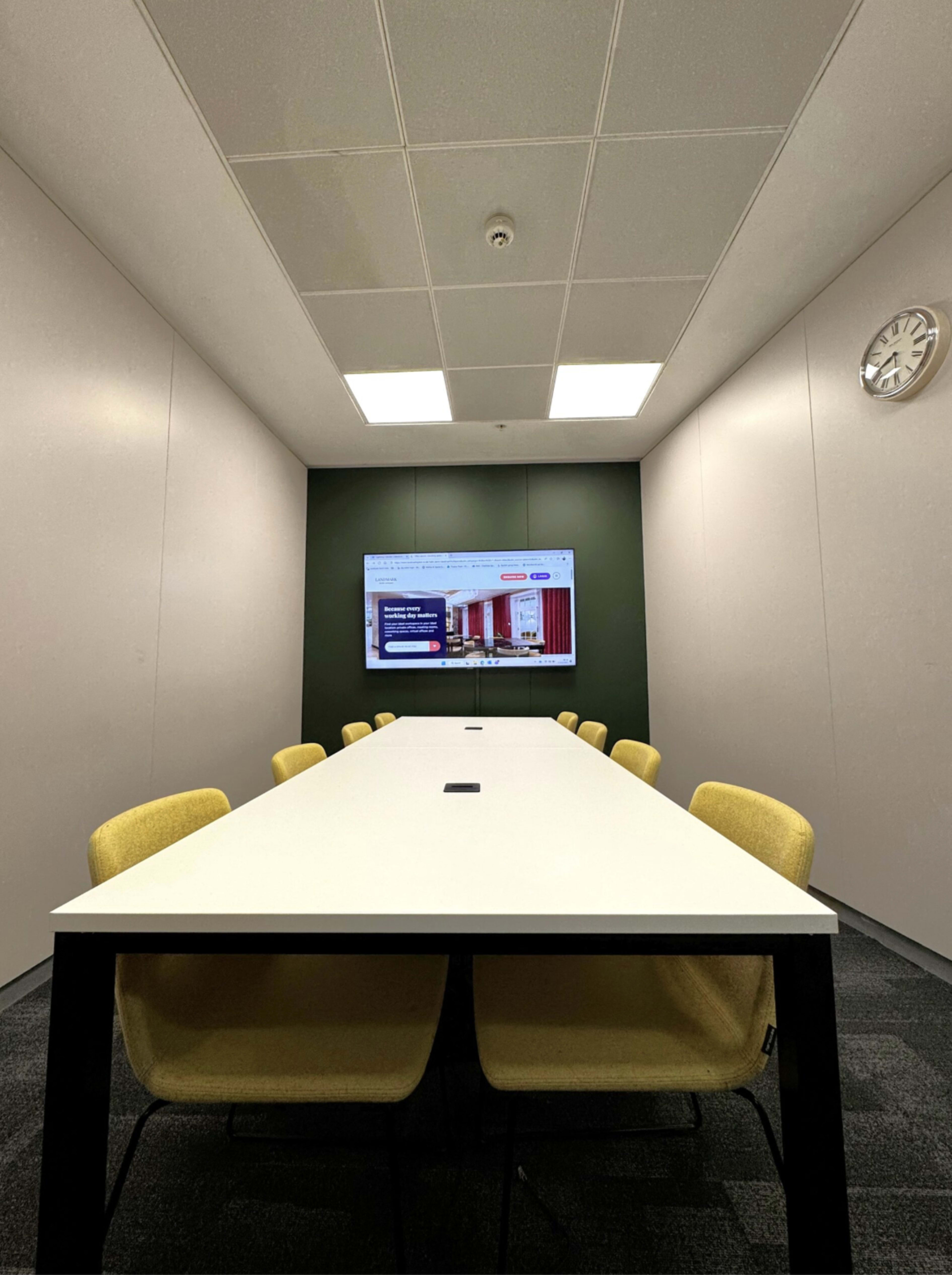 A conference room features a long white table surrounded by yellow chairs, with a large screen displaying a webpage on a green accent wall.