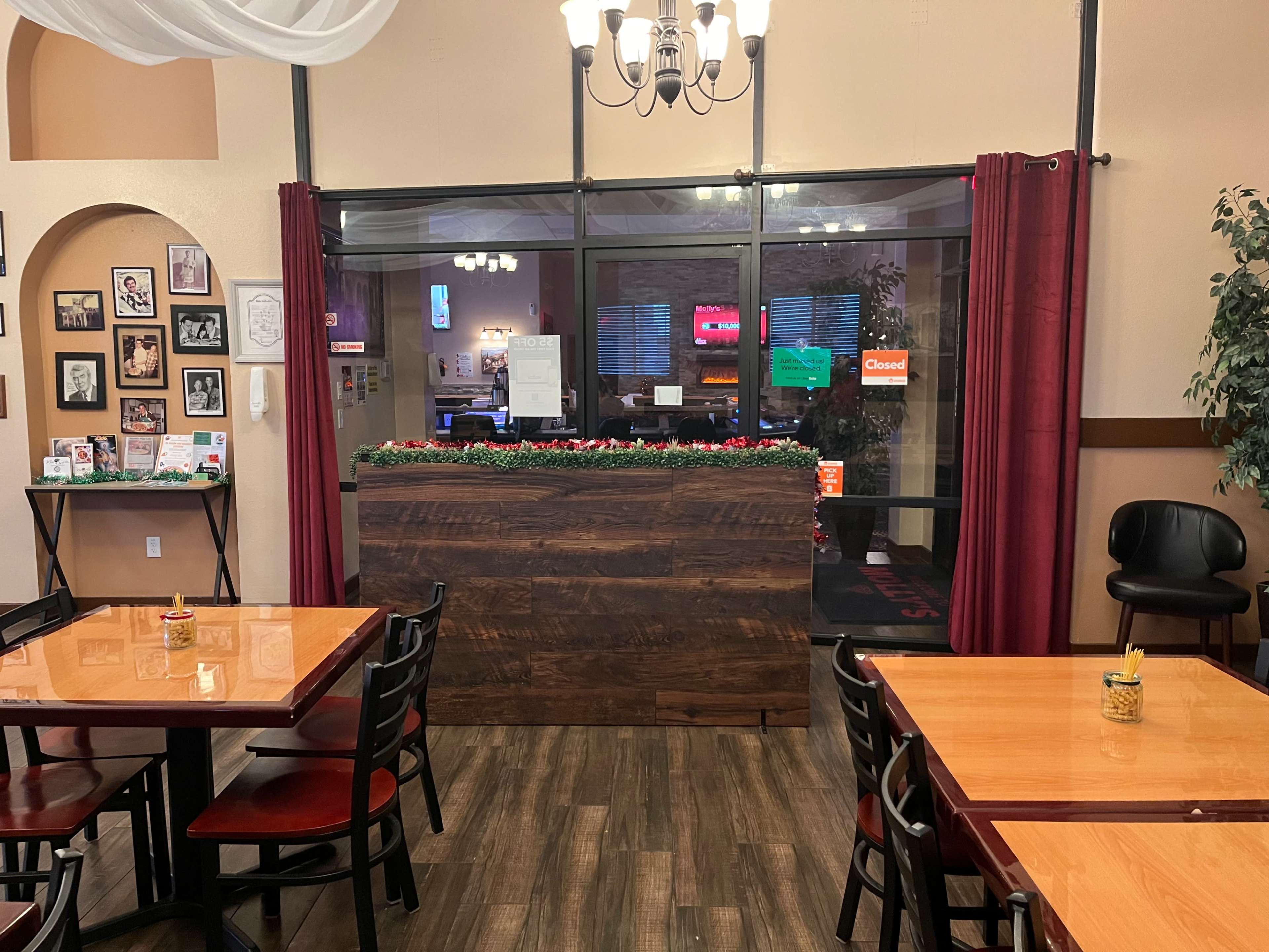 A restaurant interior with wooden tables, black chairs, and a large window displaying a closed sign, adorned with greenery along the counter.