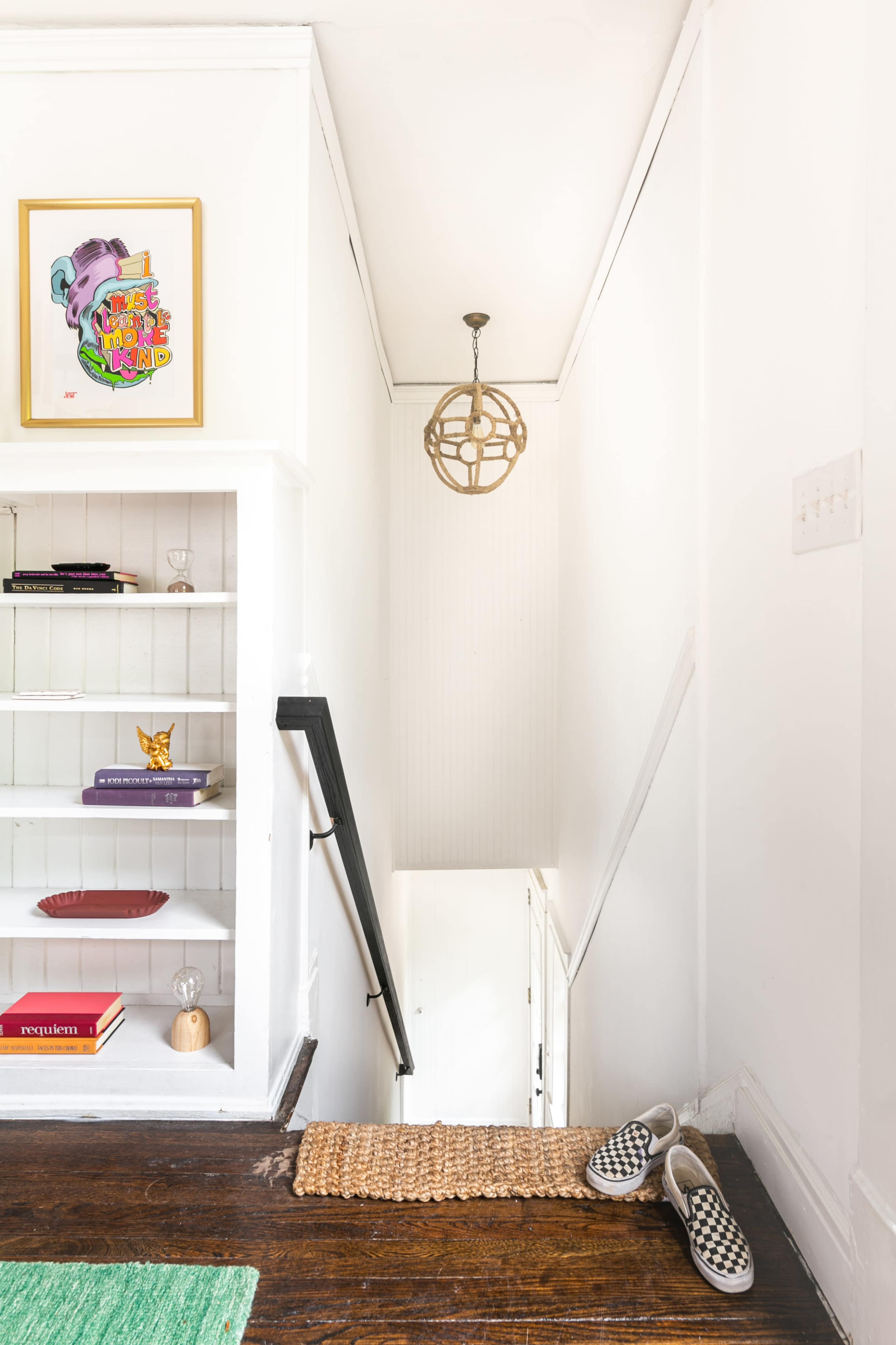 A staircase leads down to a white hallway, with a light fixture hanging above and a shelf displaying various items on the left.
