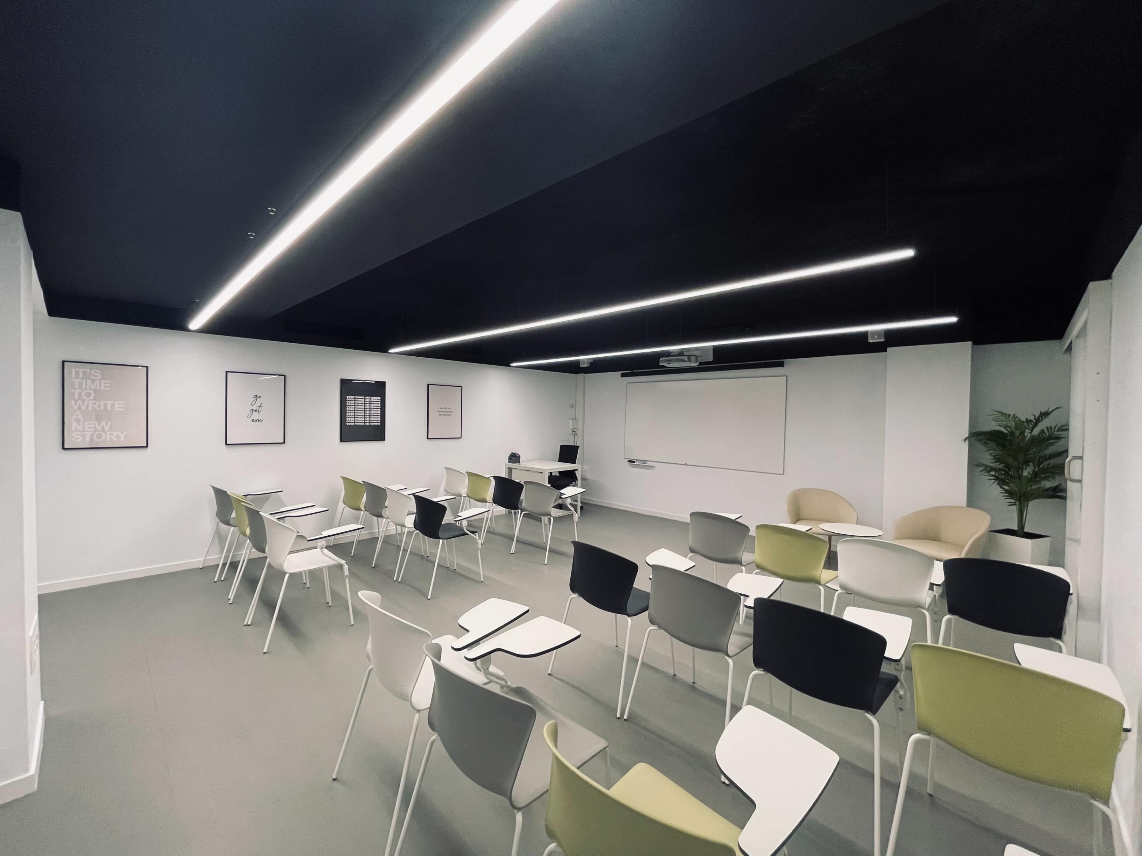 A modern classroom is arranged with rows of chairs and a whiteboard at the front, illuminated by linear ceiling lights.