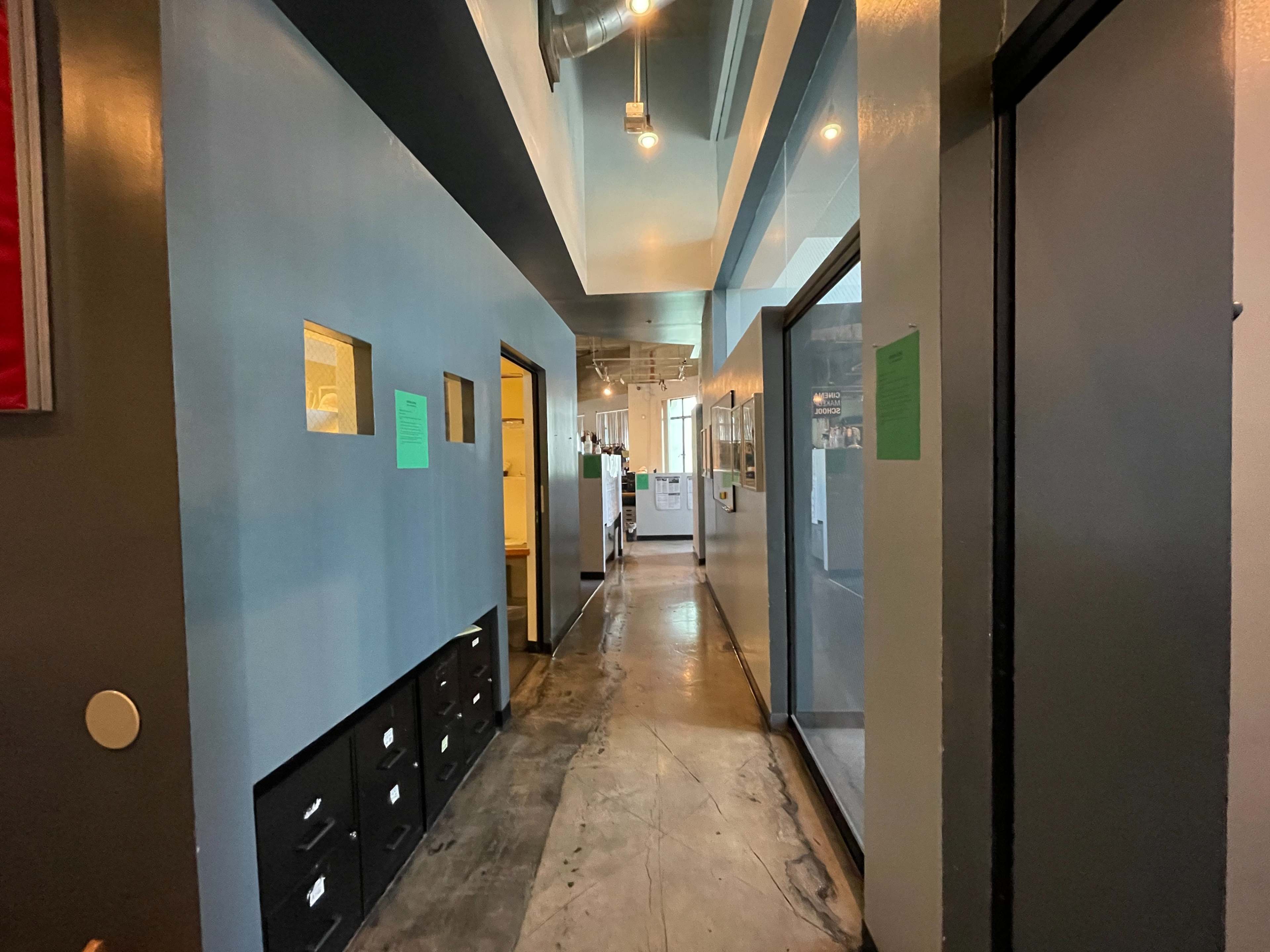 The image shows a narrow hallway with blue walls, lit by overhead lights, leading to doors on both sides and an open area at the end.