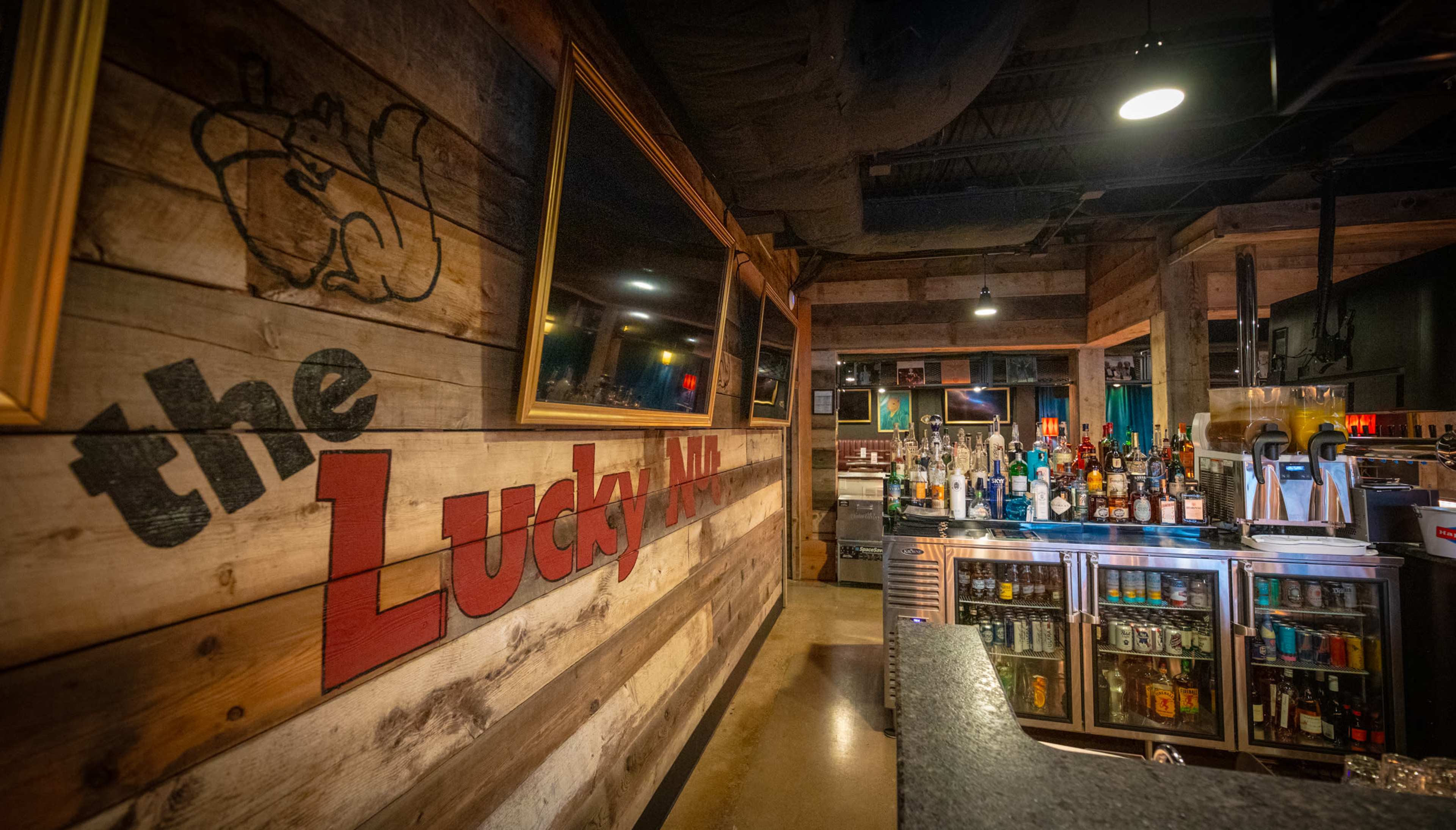 The image shows a bar interior with rustic wooden walls, featuring a sign that reads "the Lucky," and a well-stocked bar counter with various bottles displayed.