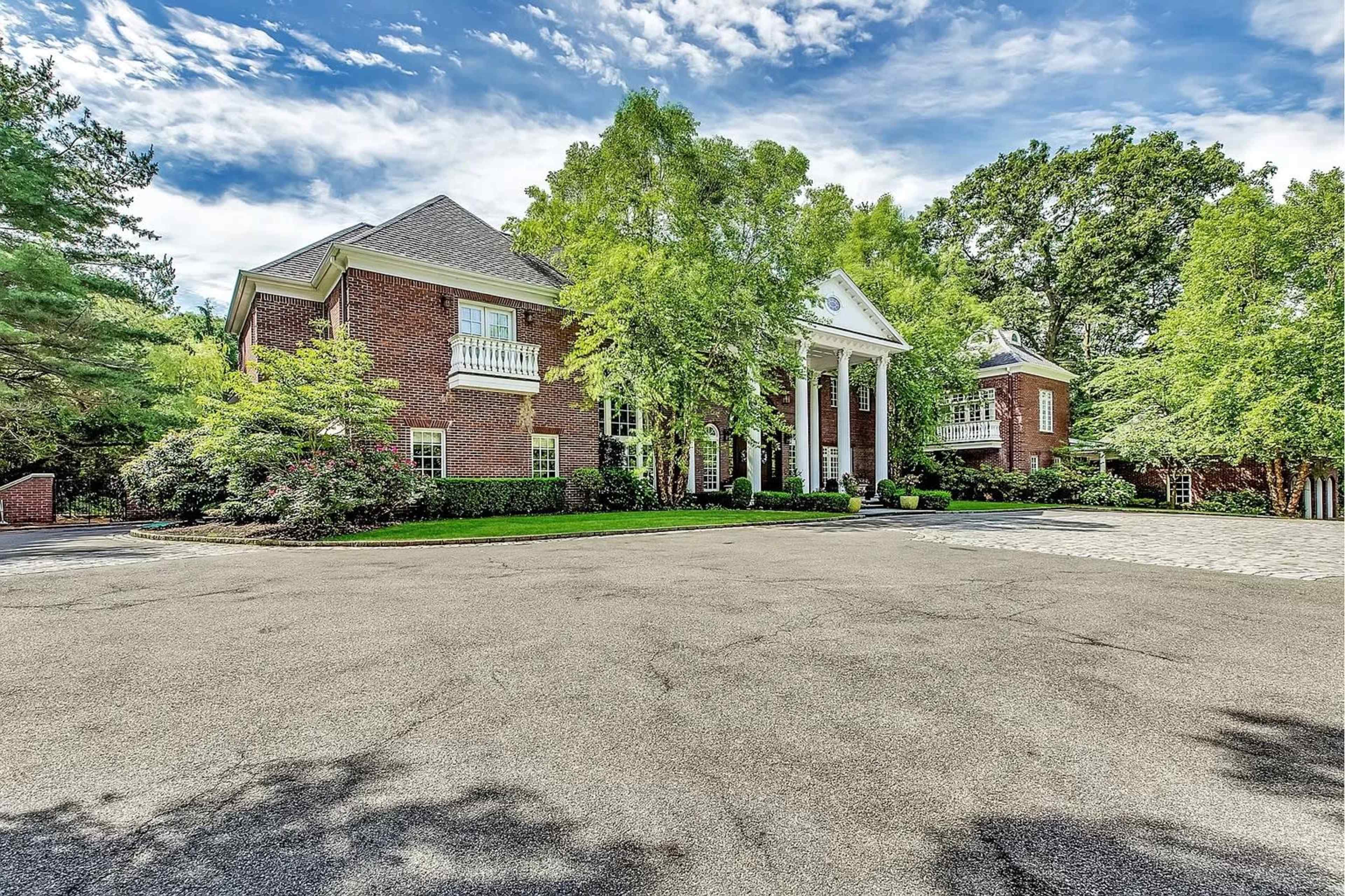 A large brick mansion with white columns, surrounded by lush landscaping and a paved driveway.